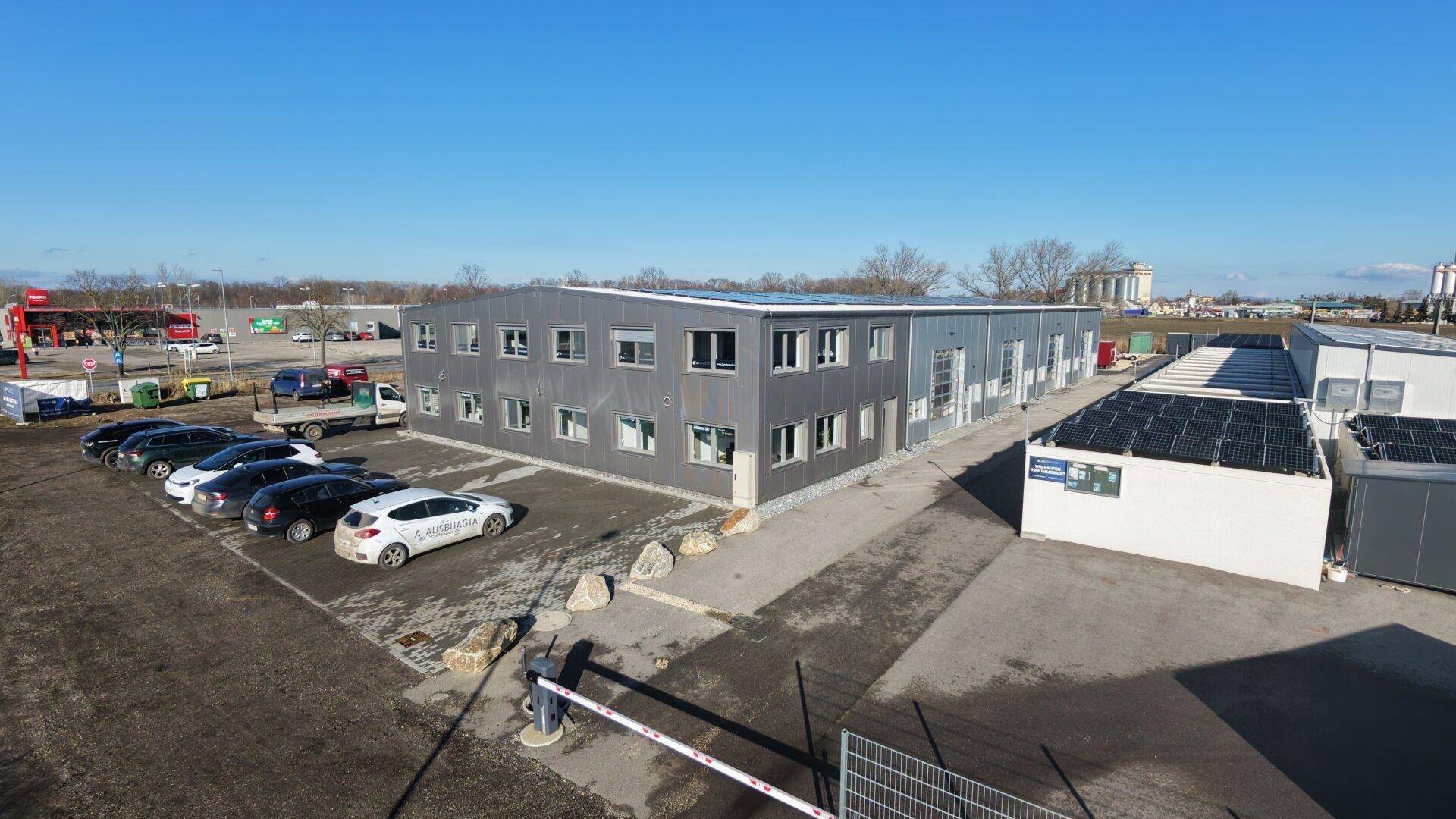 Gesamtansicht des Gewerbekomplexes mit Hauptgebäude, Nebengebäude und großem Parkplatz unter blauem Himmel.