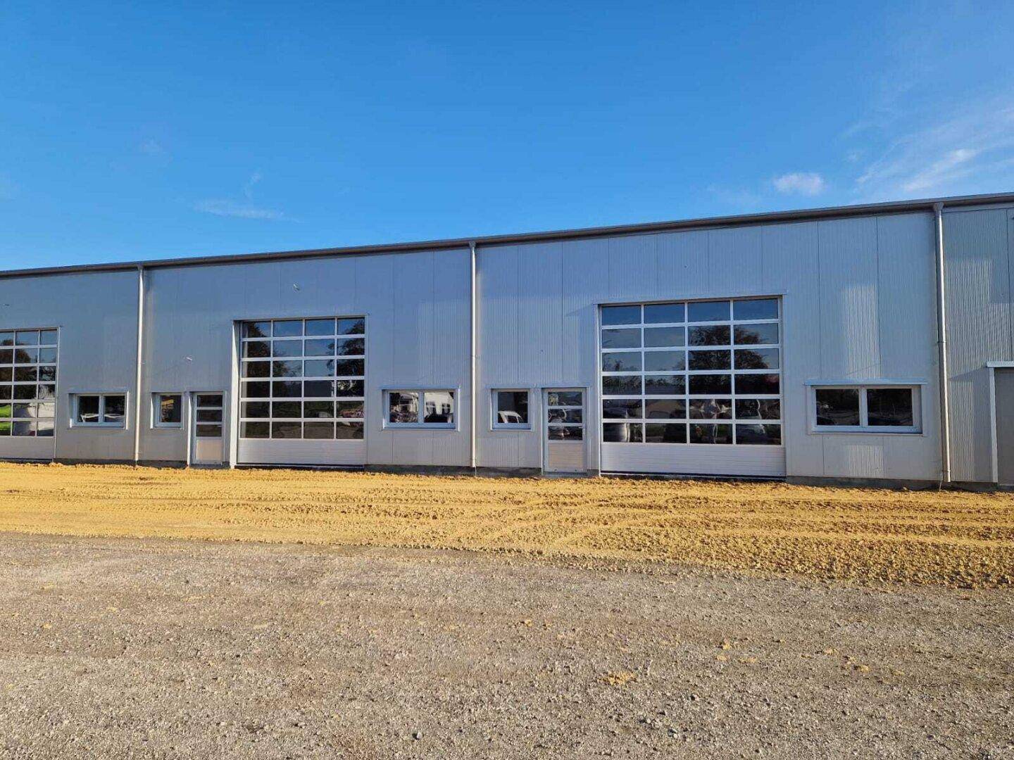 Nahaufnahme der Fassade des Gewerbegebäudes mit zwei großen, verglasten Garagentoren und unfertigem Vorplatz.