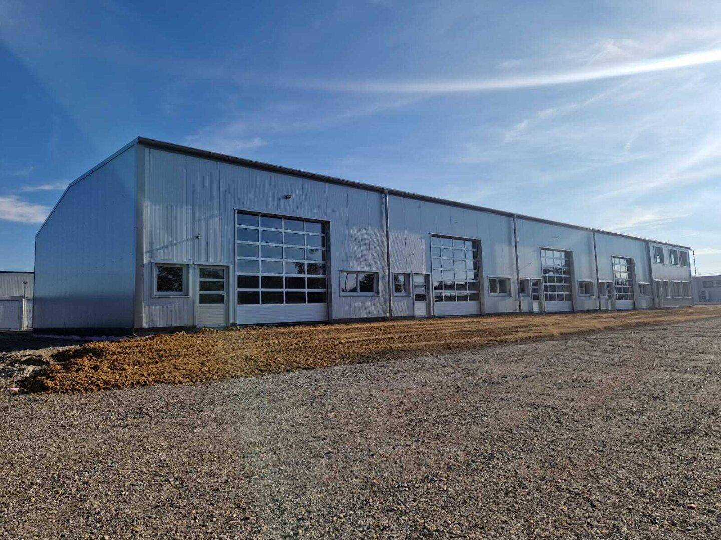Ansicht der modernen Gewerbehalle mit großen Garagentoren und einer unbefestigten Fläche im Vordergrund.