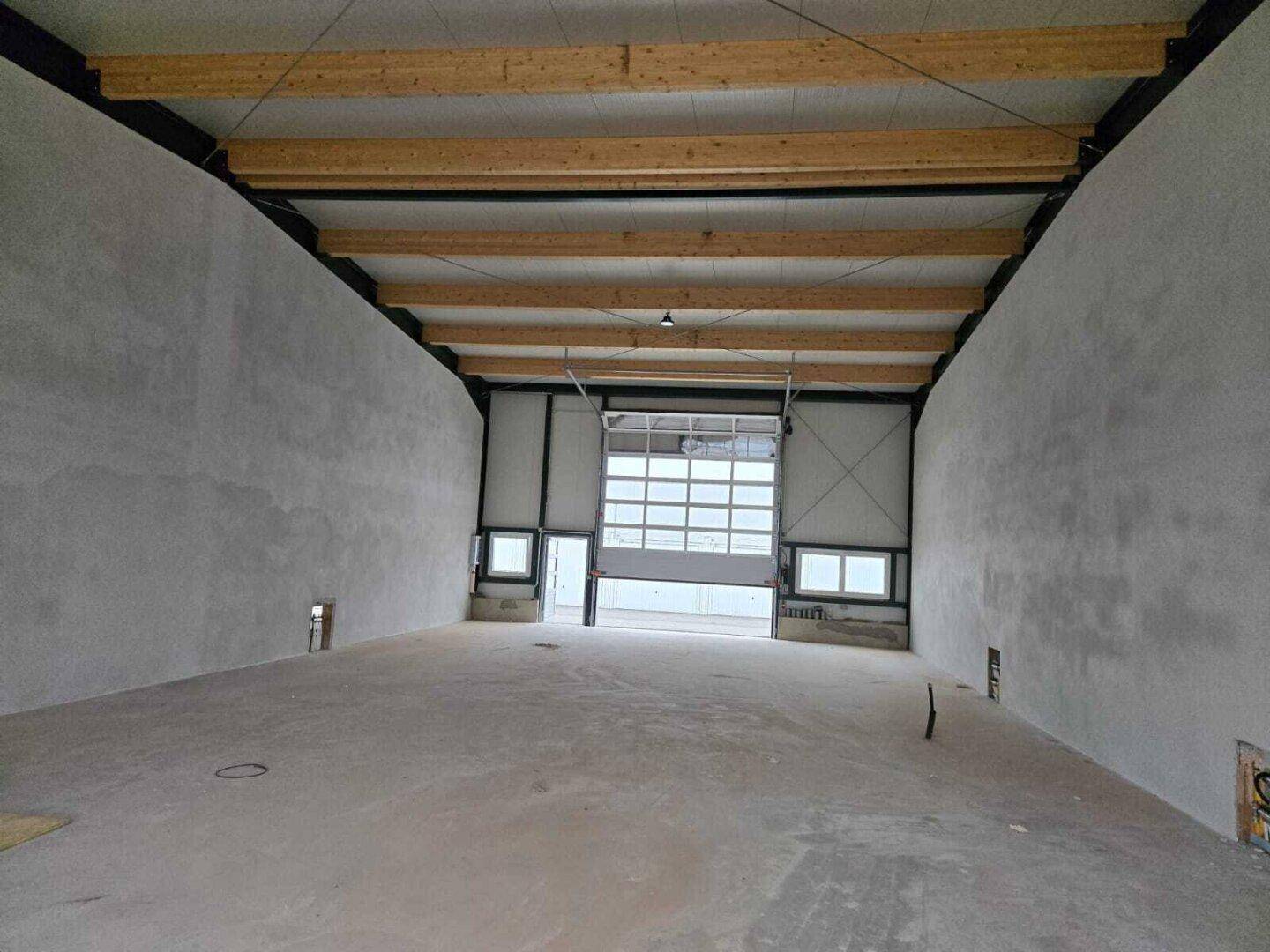 Geräumige, leere Lagerhalle mit hohen Decken, sichtbaren Holzbalken und einem großen Rolltor.