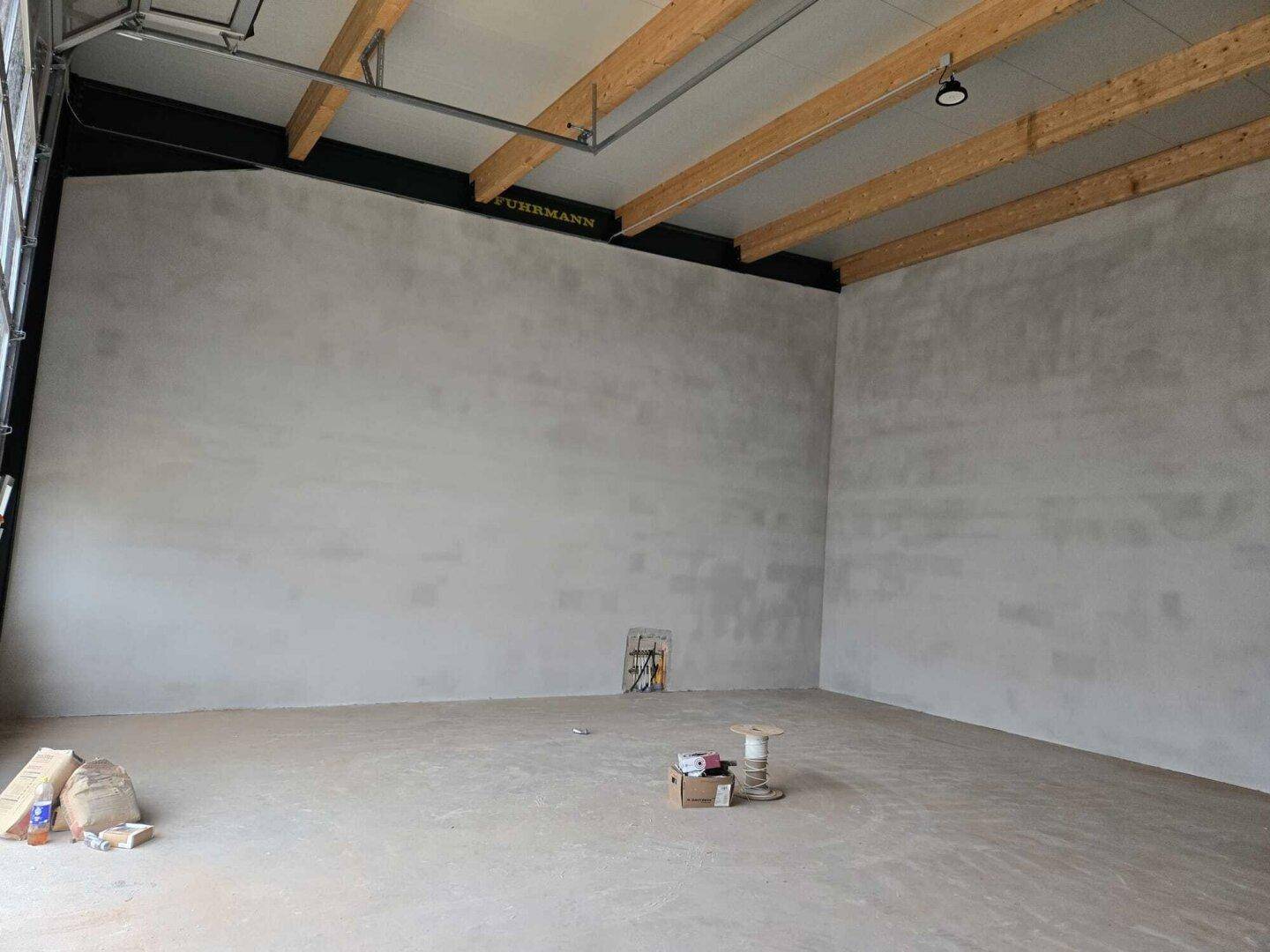 Innenansicht einer Lagerhalle mit hohen Decken, Holzbalken und einer großen, ungestalteten Betonwand.