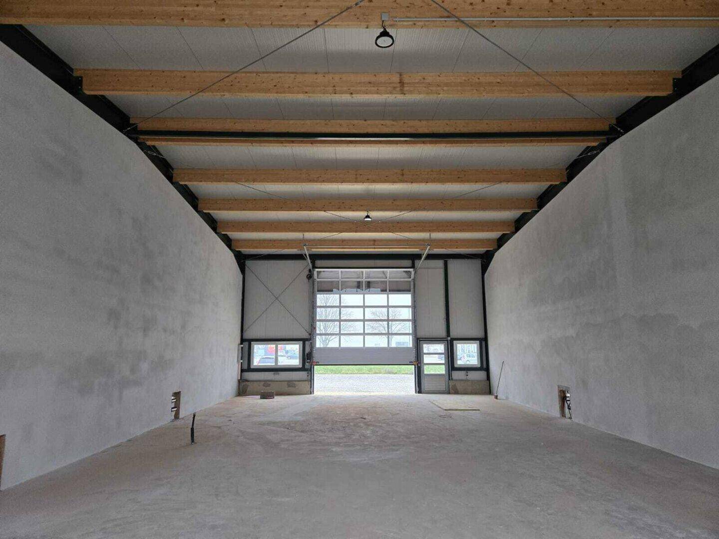 Leere Industriehalle mit hohen Decken, sichtbaren Holzbalken und einem großen, verglasten Sektionaltor.
