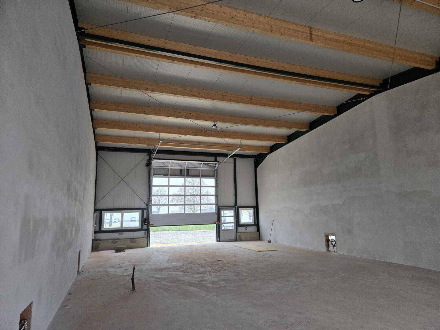 Innenansicht einer neuen Lagerhalle mit hohen Decken, Holzbalken und einem großen, geöffneten Sektionaltor.