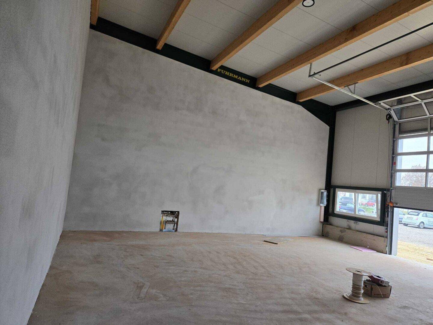 Innenraum einer Lagerhalle mit hohen Decken, Holzbalken und einer großen, ungestalteten Betonwand.