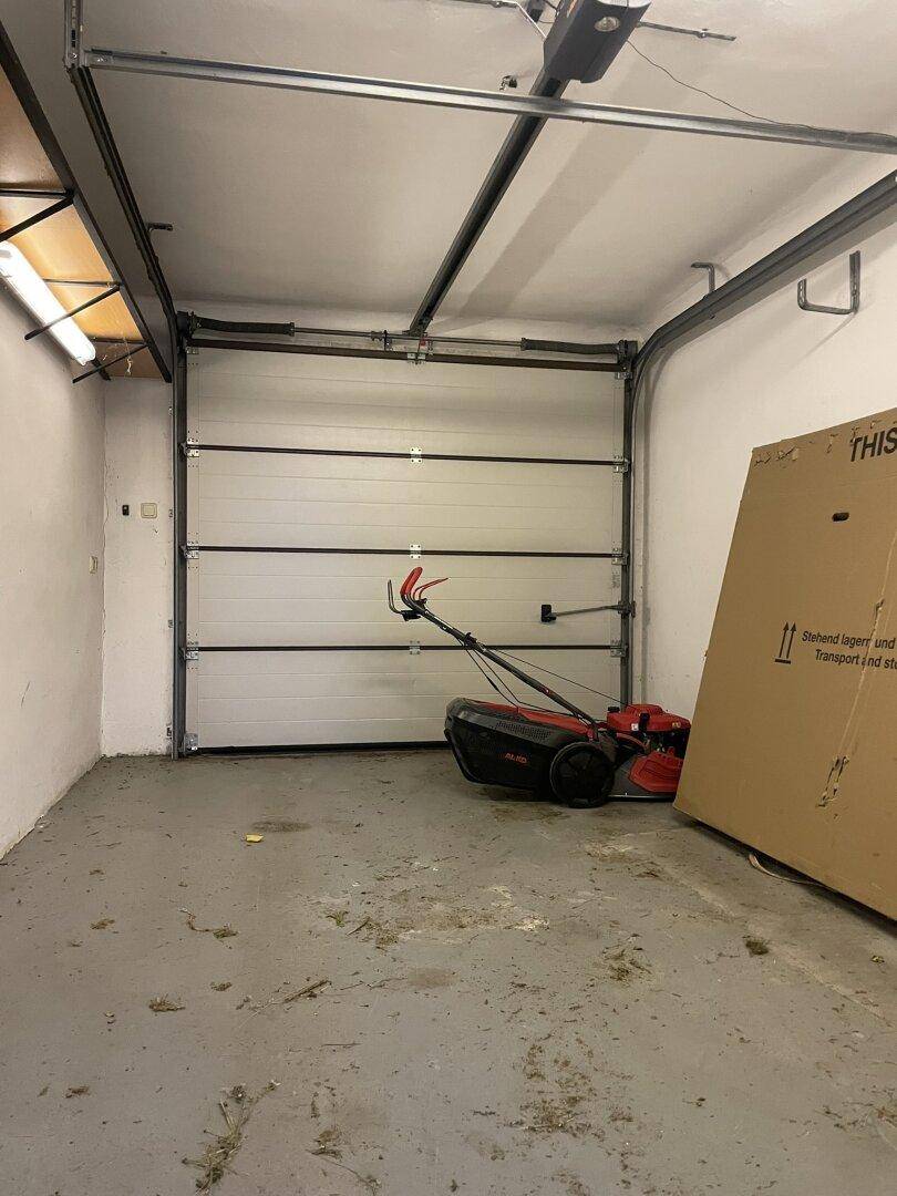 Geräumige Garage mit Sektionaltor, Betonboden und Rasenmäher.