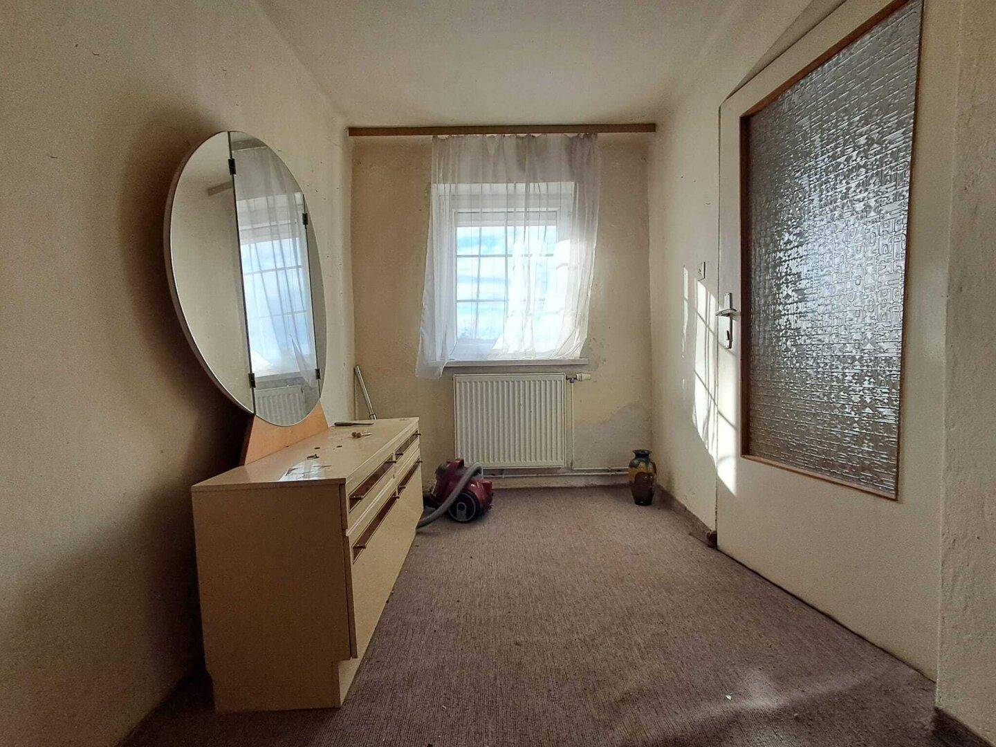 Schmaler Raum mit Teppichboden, Kommode mit Spiegel und einem Fenster für natürliches Licht.
