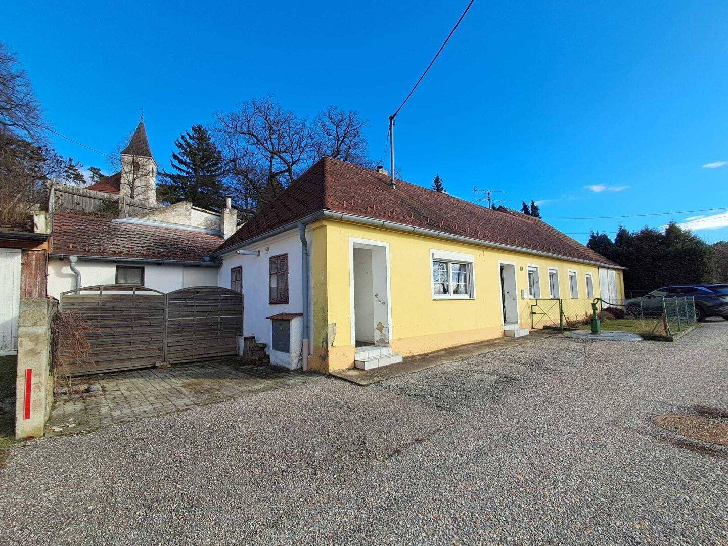 Seitenansicht eines gelben Hauses mit Kiesfläche und Blick auf eine Kirche im Hintergrund.