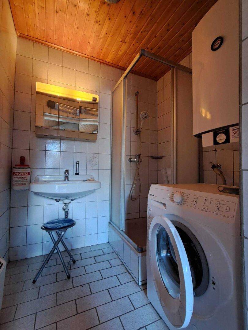 Kleines Badezimmer mit Dusche, Waschbecken und Waschmaschine unter Holzdecke.