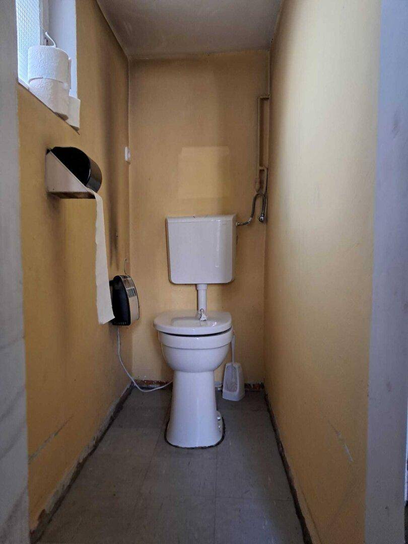 Kleines WC mit Toilette, Fenster und Wandspender für Papier in gelbem Raum.