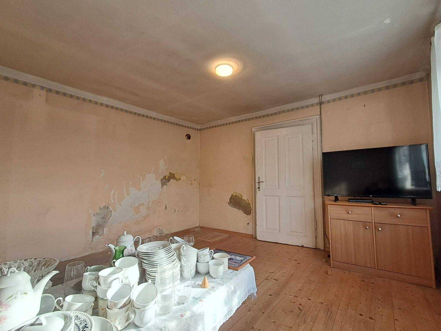 Wohnraum mit Holzdielenboden, weißer Tür und einem Fernseher auf einem Holzschrank.