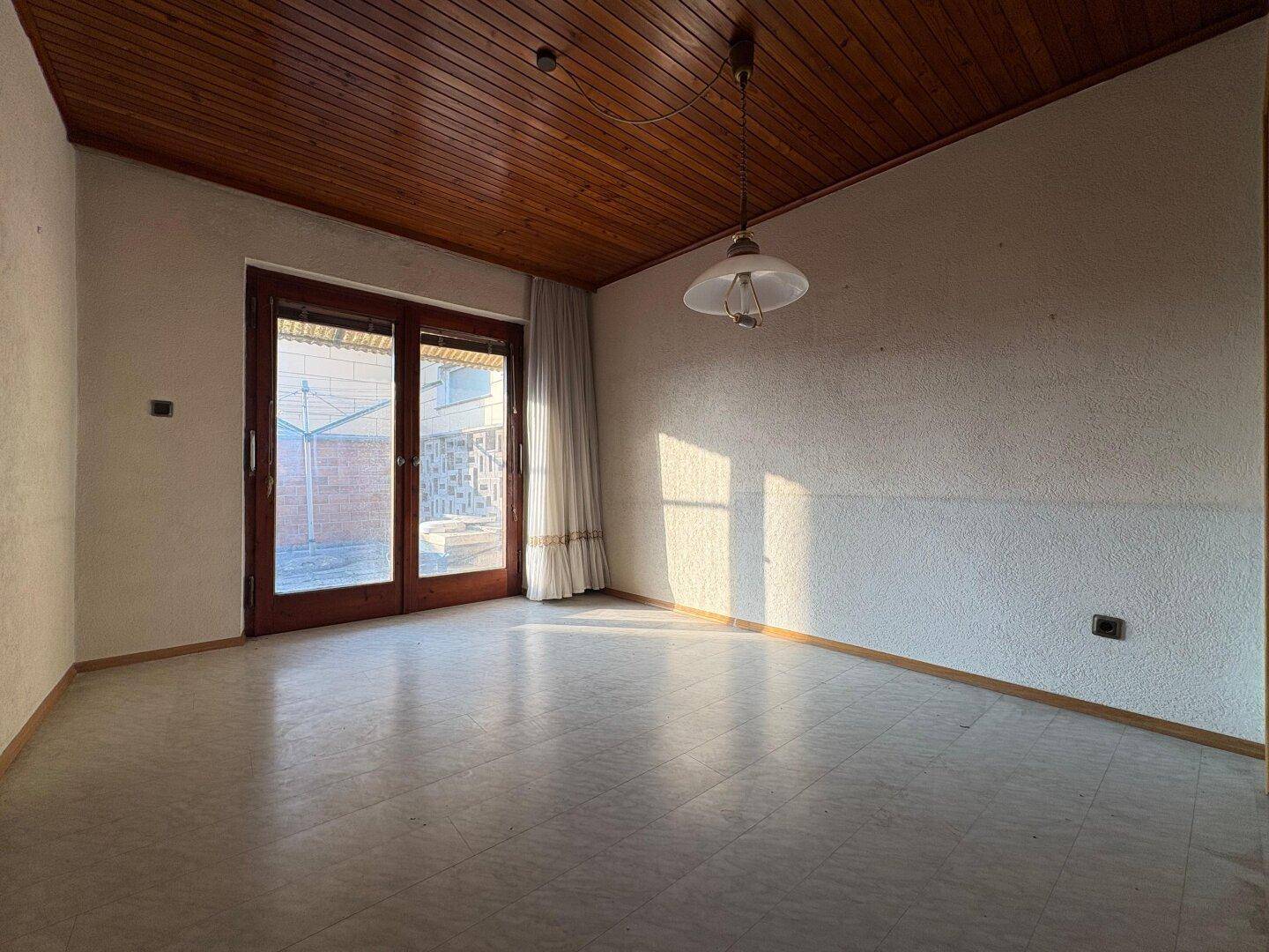 Raum mit Holzdecke und Fliesenboden, der durch eine Glastür Zugang zum Außenbereich bietet.