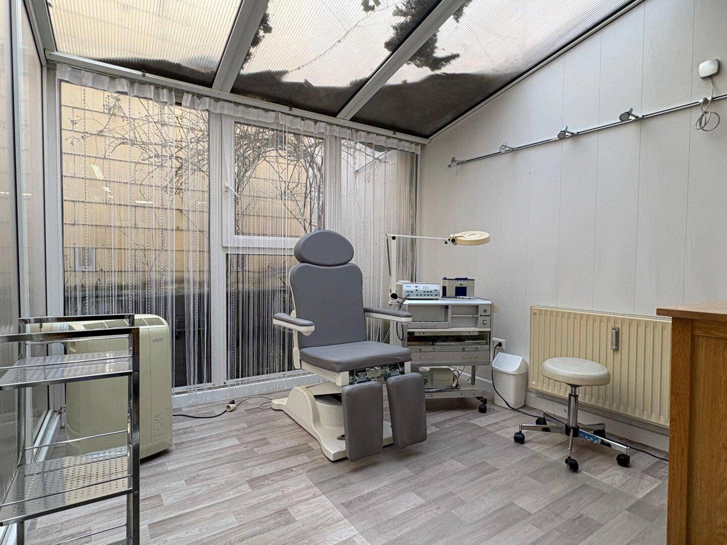 Heller Wintergarten mit Behandlungsliege und viel Tageslicht, ideal für Wellness oder Therapien.