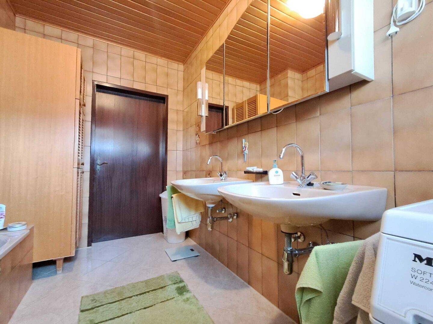 Badezimmer mit Doppelwaschbecken, Spiegelschrank und braunen Fliesen, ergänzt durch eine Holzdecke.
