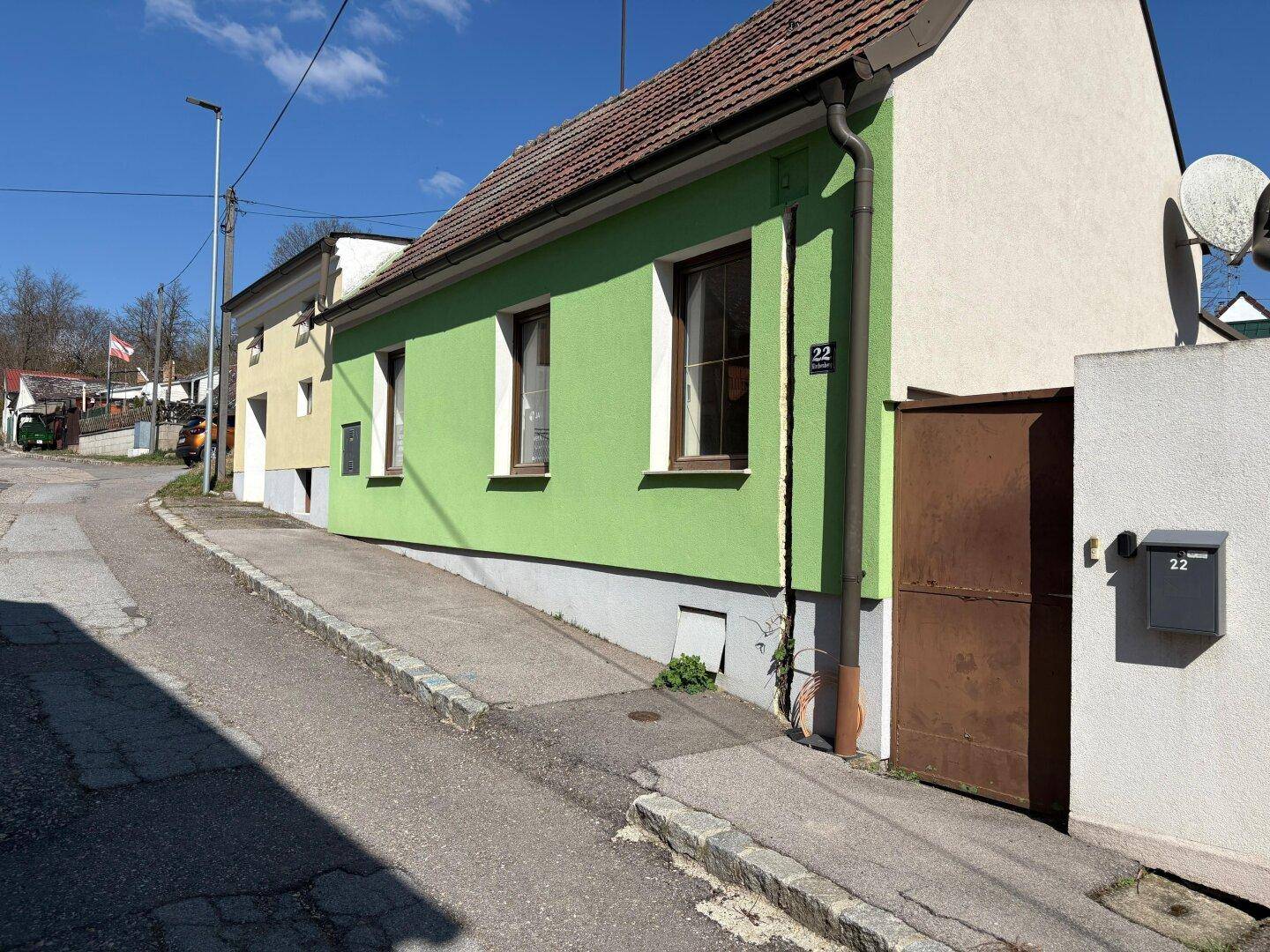 Grünes Haus mit braunen Fenstern und Haustür an einer leicht abschüssigen Straße.