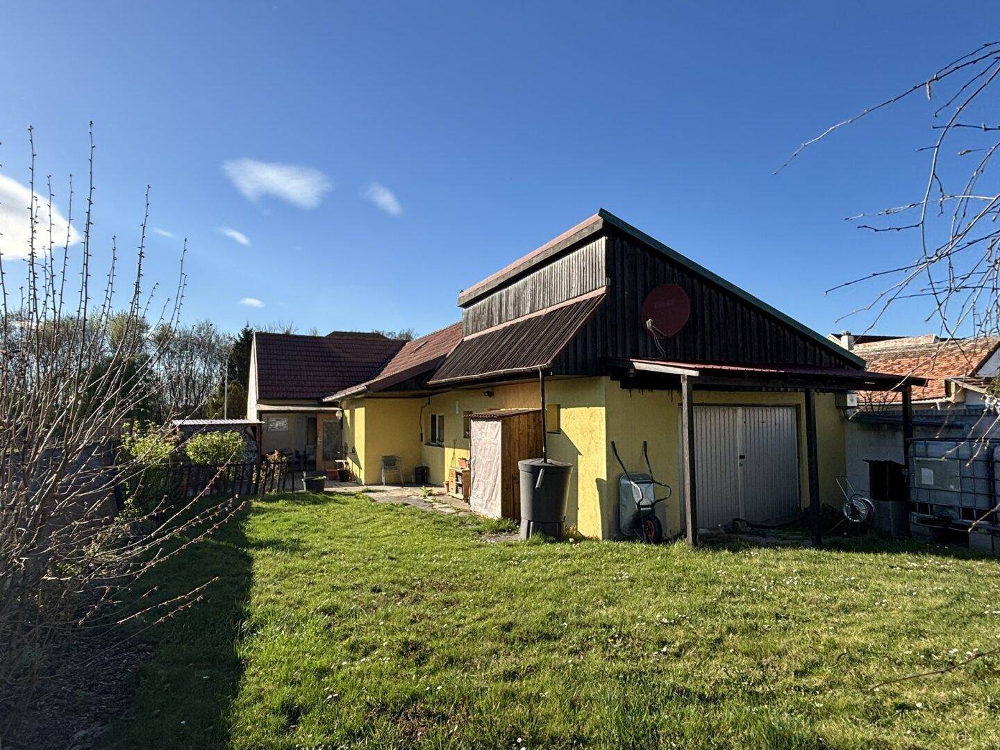 Rückansicht des Hauses mit Satteldach und Holzverkleidung, umgeben von einem grünen Garten unter blauem Himmel.