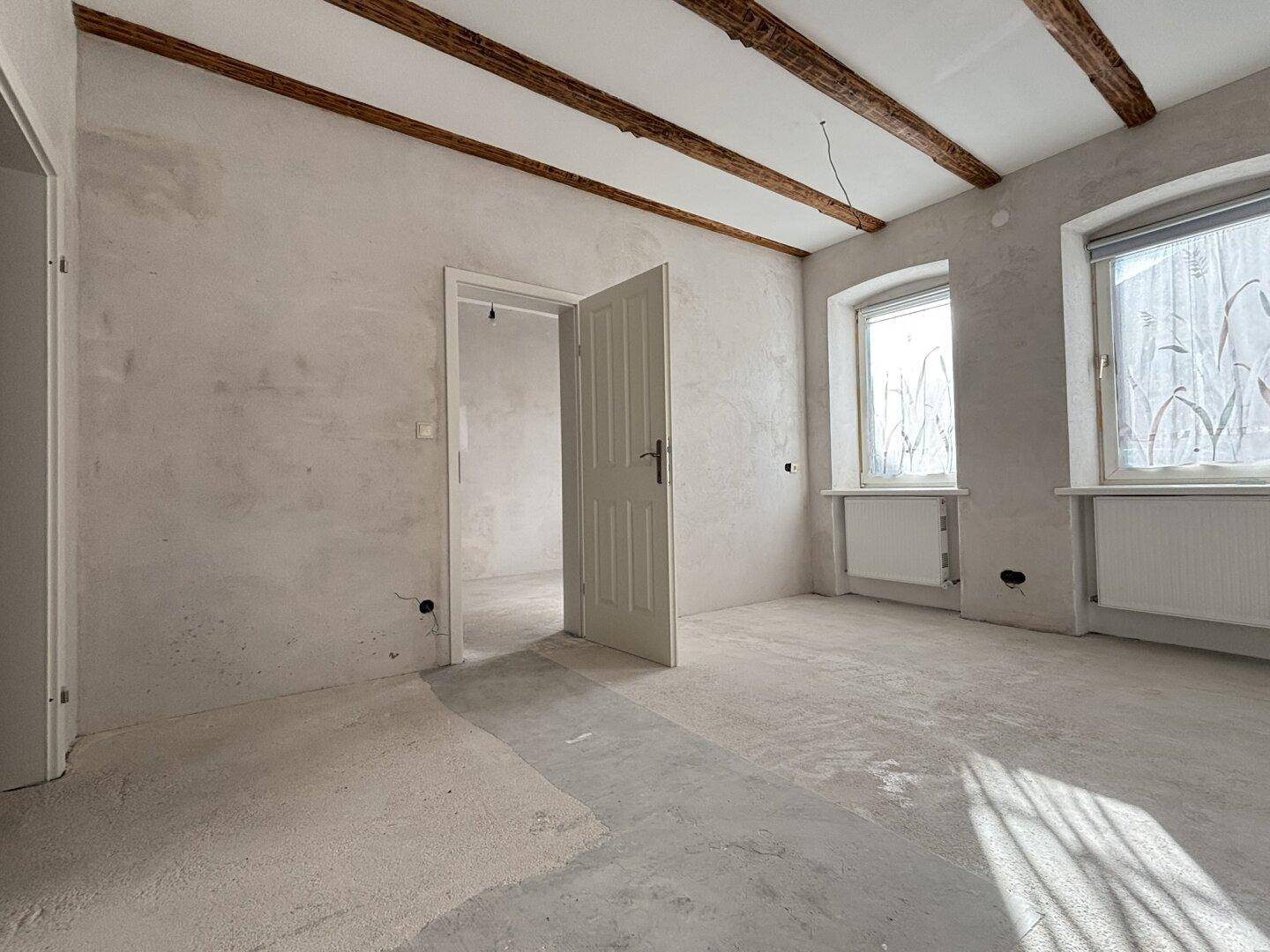 Geräumiger Raum im Rohbau mit freiliegenden Holzbalken, Betonboden und großen Fenstern.