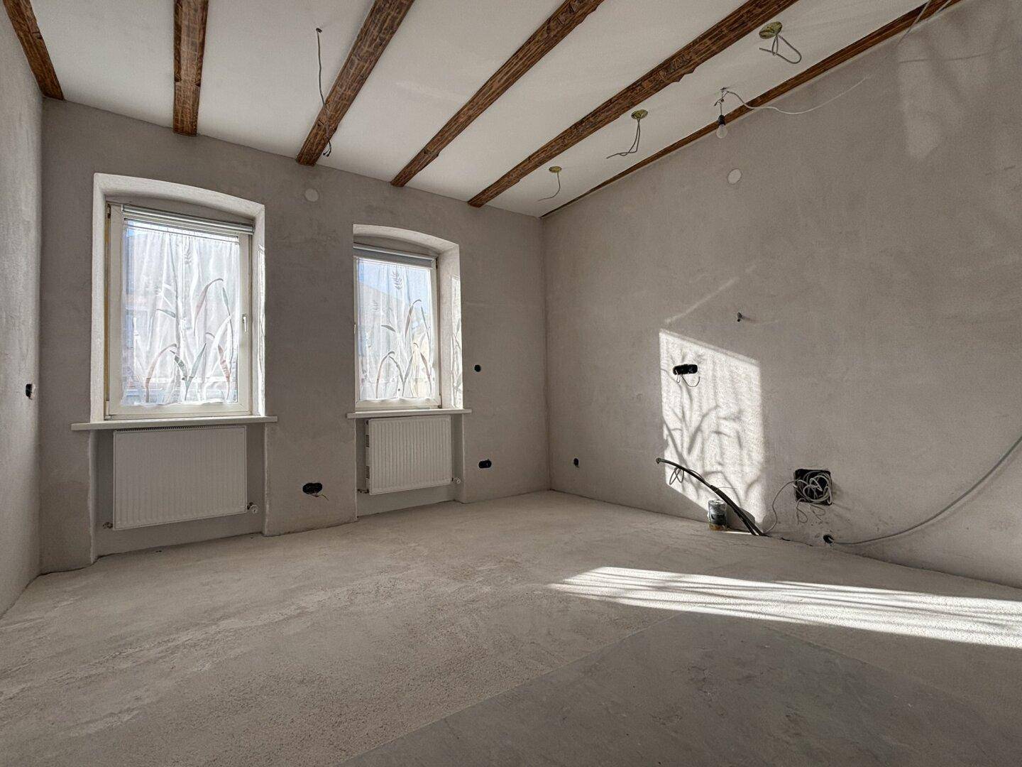 Heller Raum im Rohbau mit sichtbaren Deckenbalken, Betonboden und viel Tageslicht durch die Fenster.