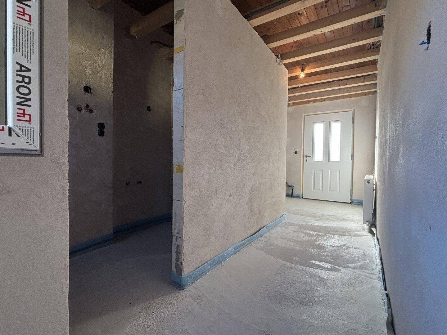 Flur im Rohbau mit sichtbaren Deckenbalken und neuer Eingangstür, bereit für den Innenausbau.