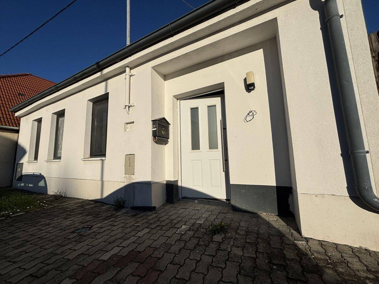 Vorderansicht des Hauses mit weißer Fassade, Eingangstür und gepflastertem Vorplatz.