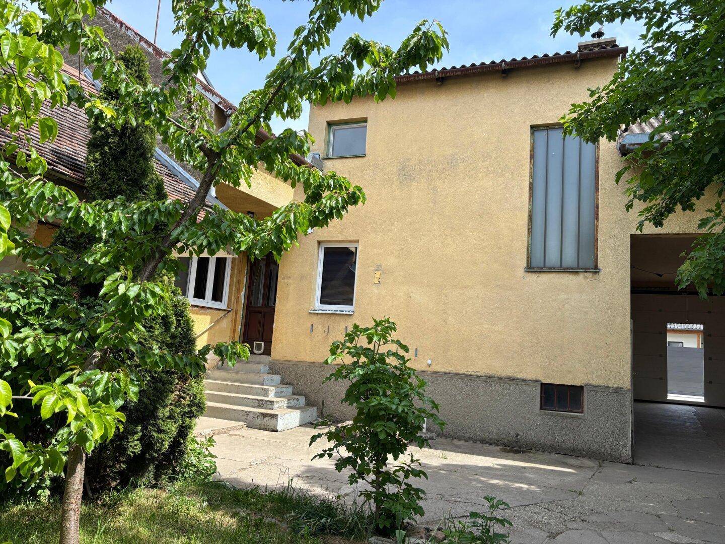 Fassade eines älteren Gebäudes mit gelbem Putz und verschiedenen Fenstern, umgeben von Bäumen.