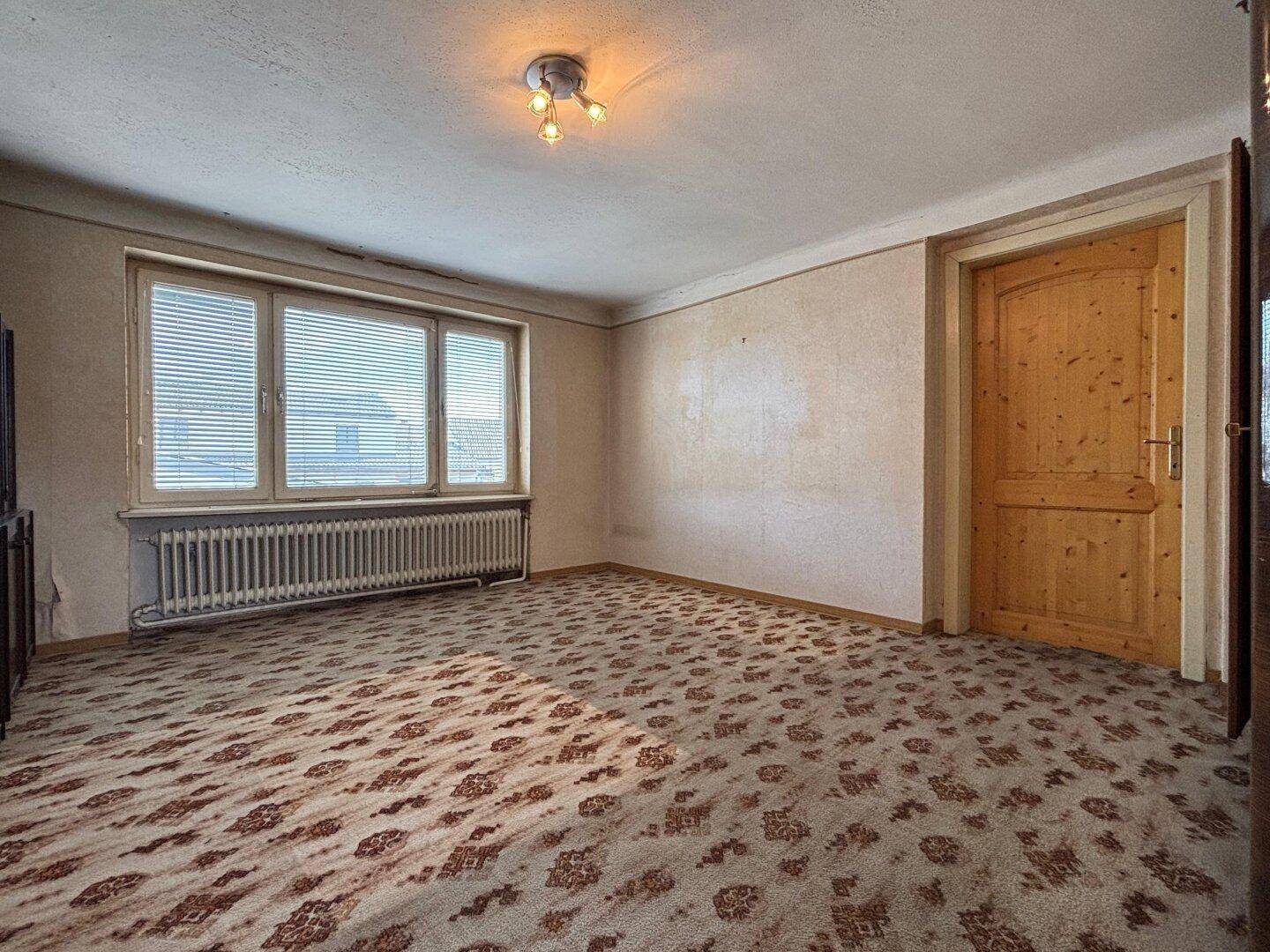 Ein Zimmer mit gemustertem Teppichboden, Fenster und Heizkörper, das eine Renovierung benötigt.