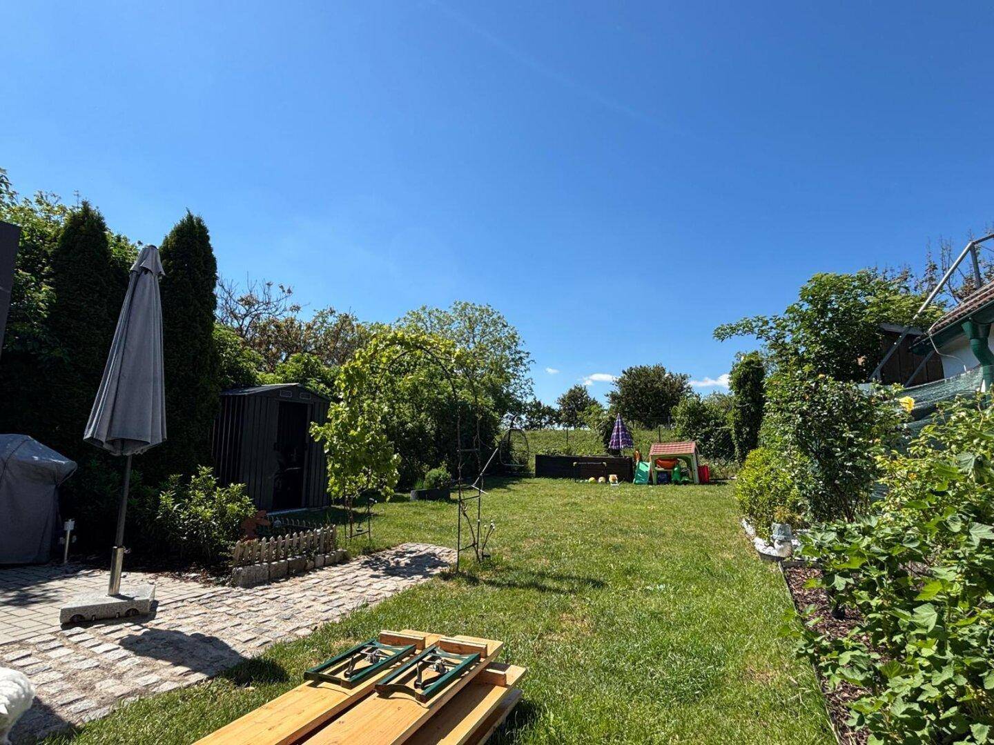 Großer, gepflegter Garten mit Rasenfläche, Bäumen und einem Gartenhaus unter blauem Himmel.