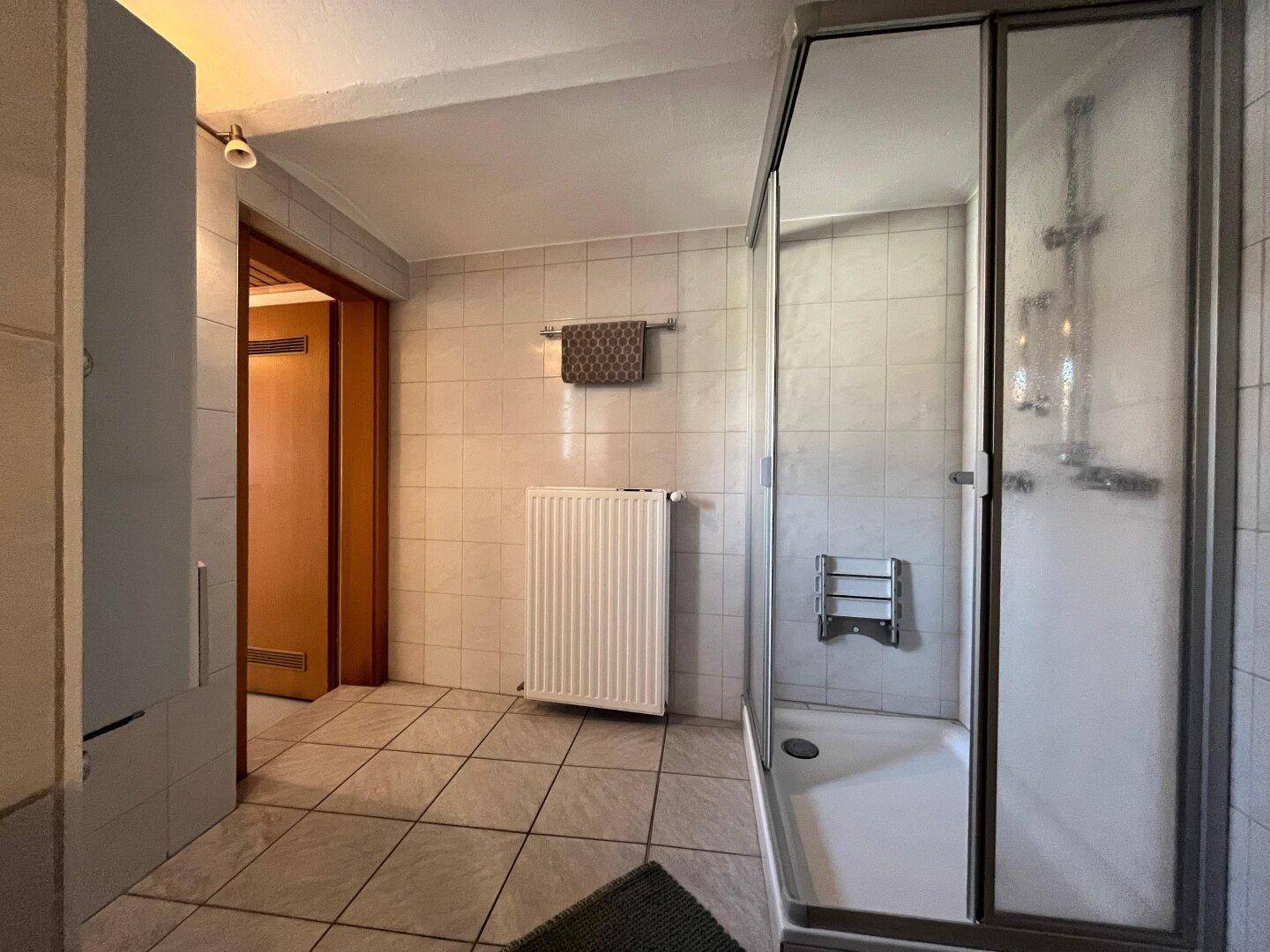 Gefliestes Badezimmer mit Duschkabine, Heizkörper und Zugang zu einem weiteren Raum.