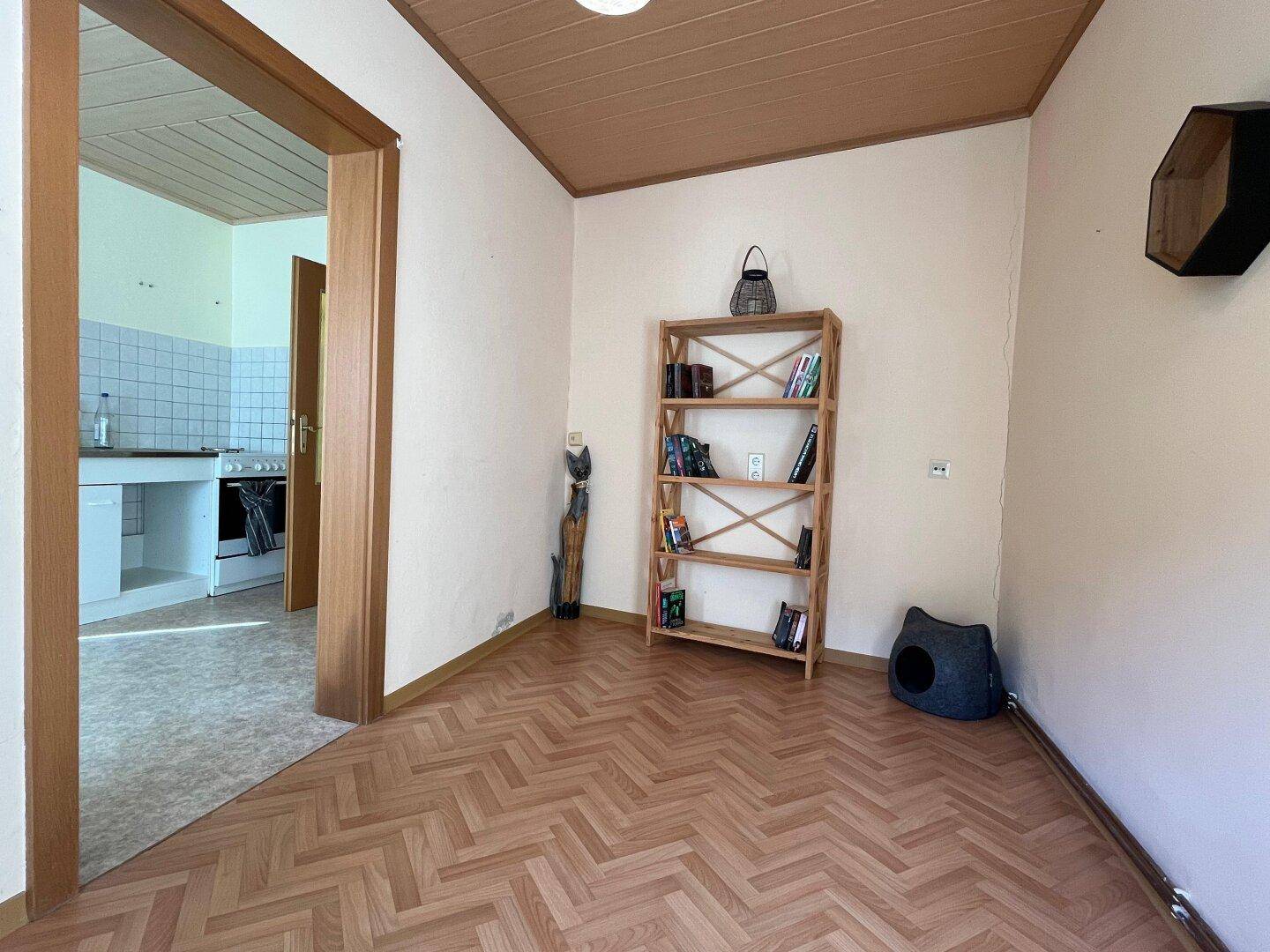Heller Raum mit Laminatboden, Bücherregal und Zugang zu einer kleinen Küchenzeile.