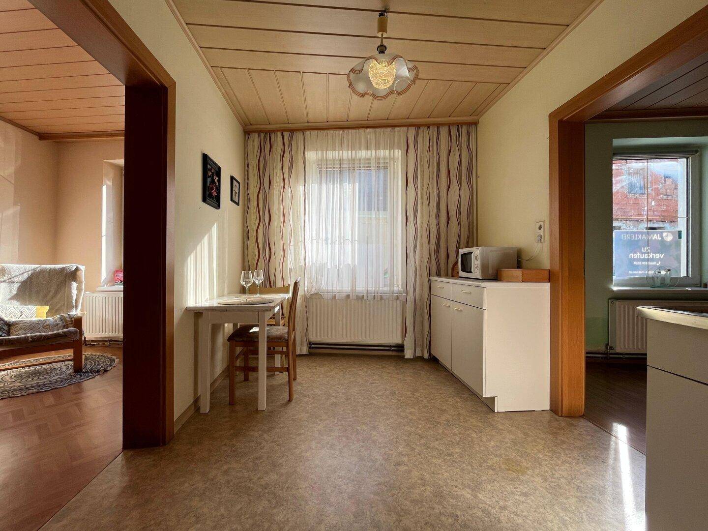 Heller Küchenbereich mit Holzbalkendecke, kleinem Esstisch und Mikrowelle am Fenster.