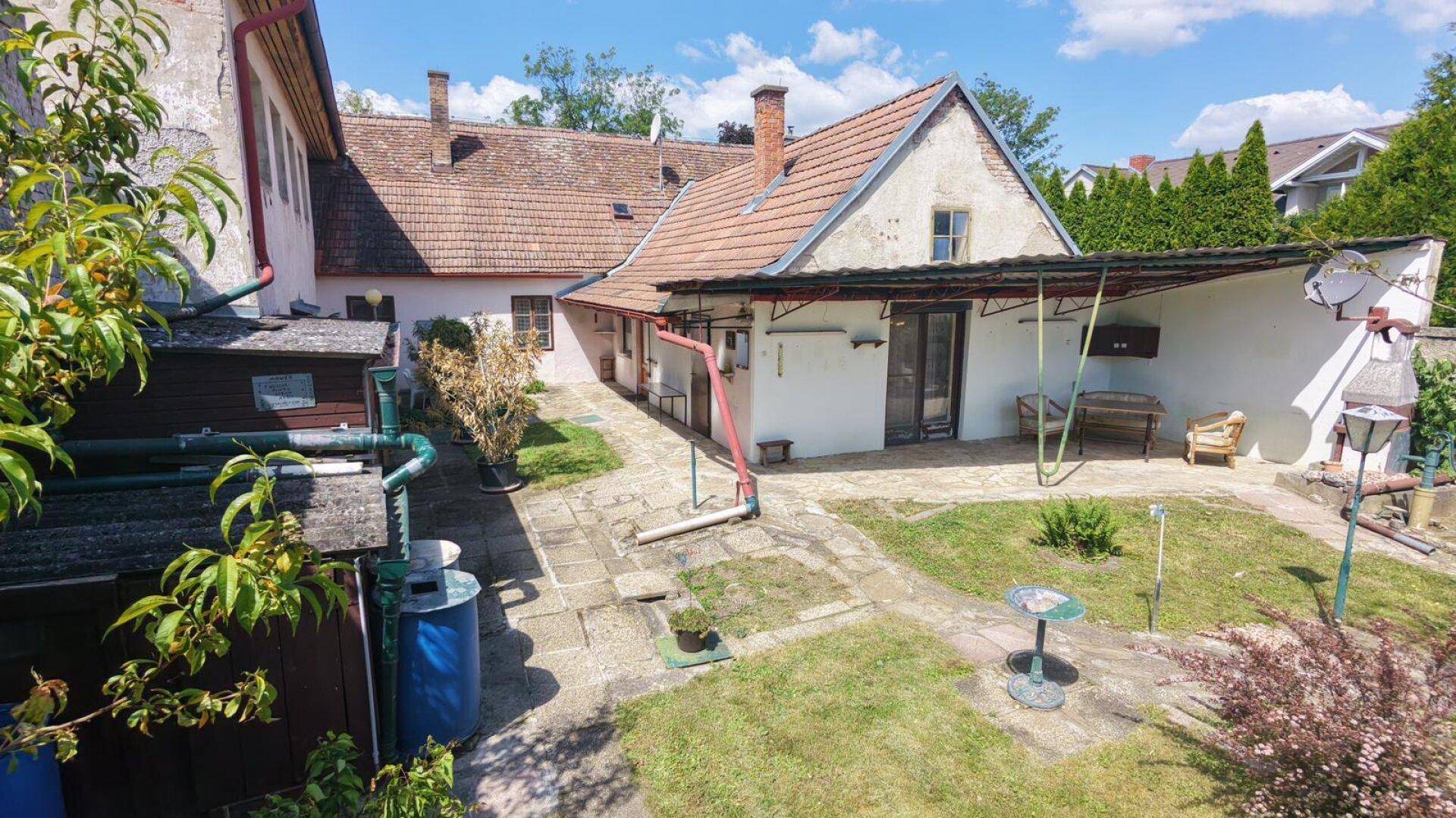 Hinterhofbereich eines älteren Gebäudes mit gepflastertem Weg, überdachtem Sitzbereich und Grünflächen.