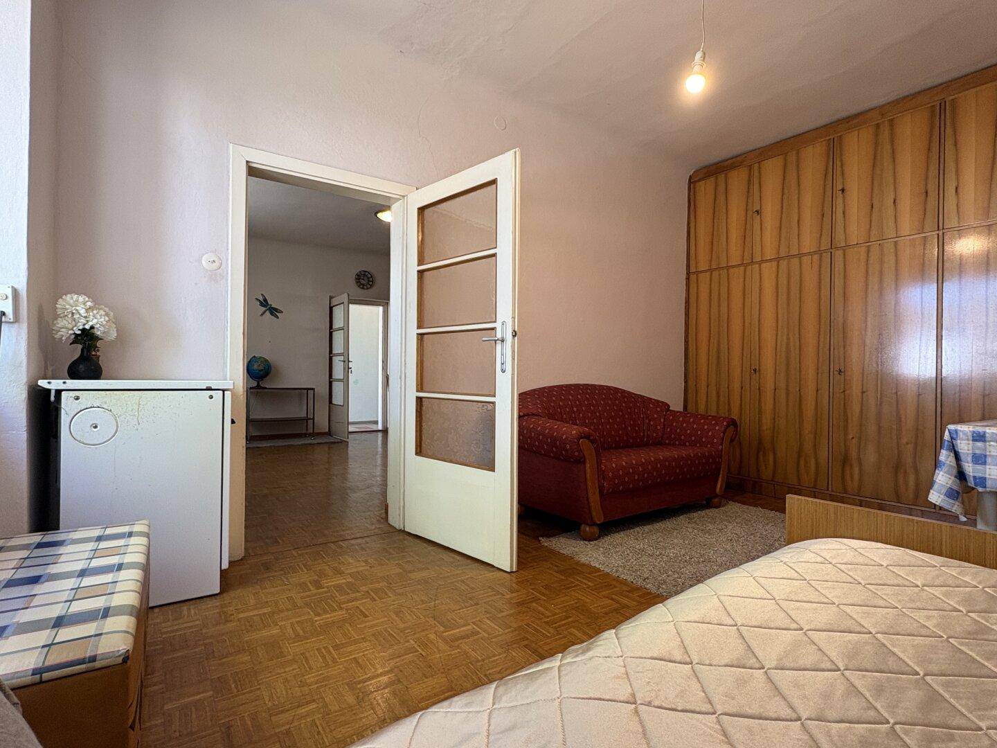 Gemütliches Schlafzimmer mit Parkettboden, rotem Sessel und Zugang zu einem weiteren Raum.