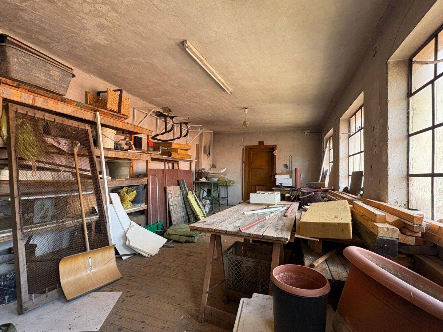 Geräumiger Werkstattraum mit großen Fenstern, Holztisch und Regalen für diverse Materialien.