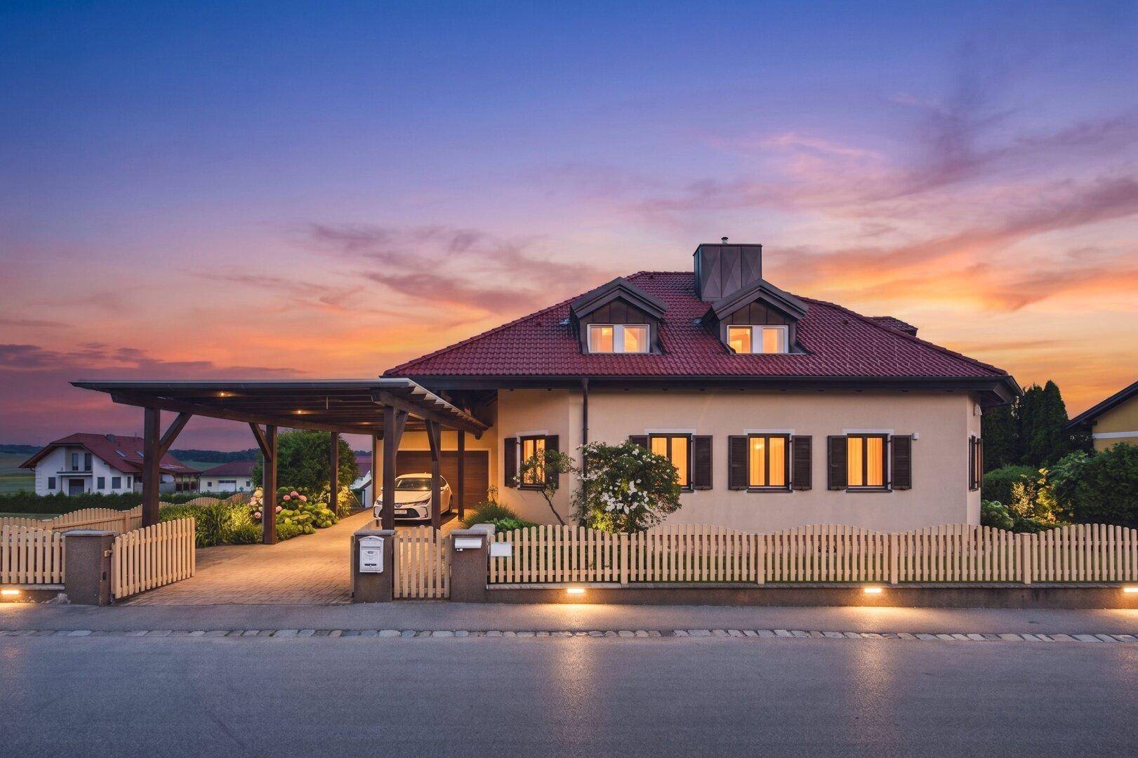 Charmantes Einfamilienhaus bei Sonnenuntergang mit Carport und gepflegtem Vorgarten, umgeben von einem Holzzaun.