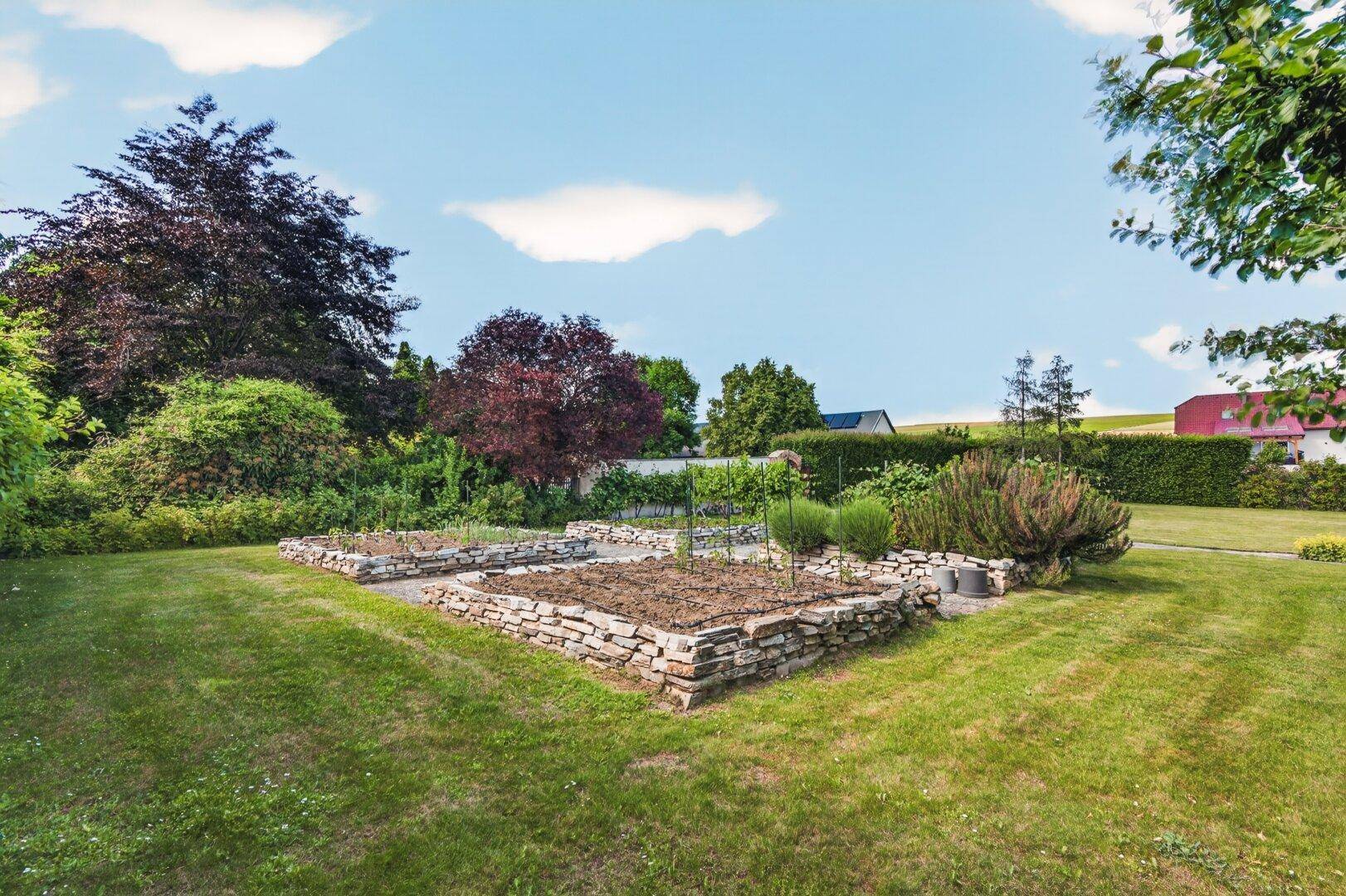 Gartenbereich mit erhöhten Steinbeeten für den Anbau, umgeben von Rasen und verschiedenen Pflanzen.
