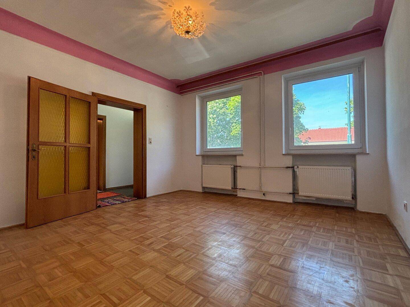 Geräumiges Zimmer mit Parkettboden, weißen Wänden und dekorativer rosa Deckenleiste.