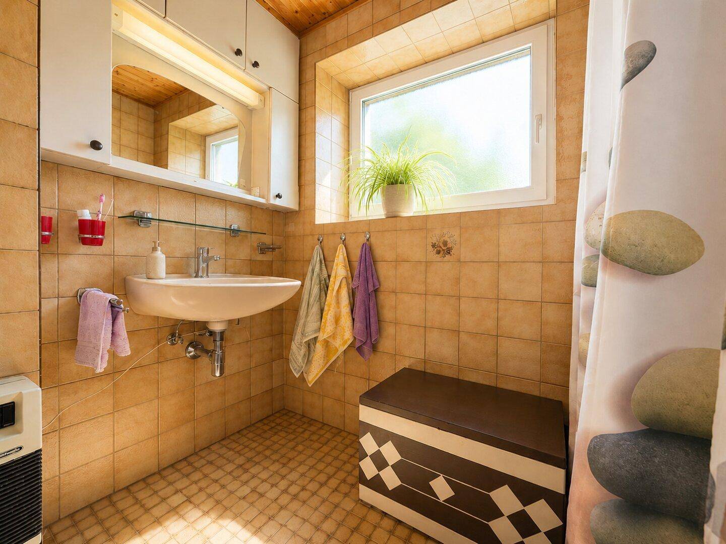 Gefliestes Badezimmer mit Waschbecken, Spiegelschrank und Fenster, in warmen Erdtönen gehalten.