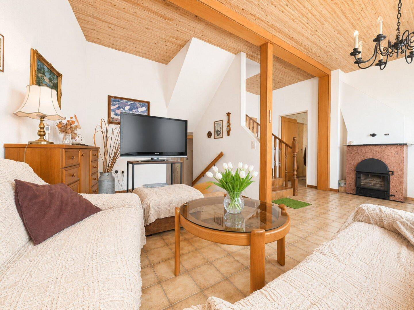 Gemütlicher Wohnbereich mit Kamin, Fliesenboden und Holzdecke, hell durch große Fenster.