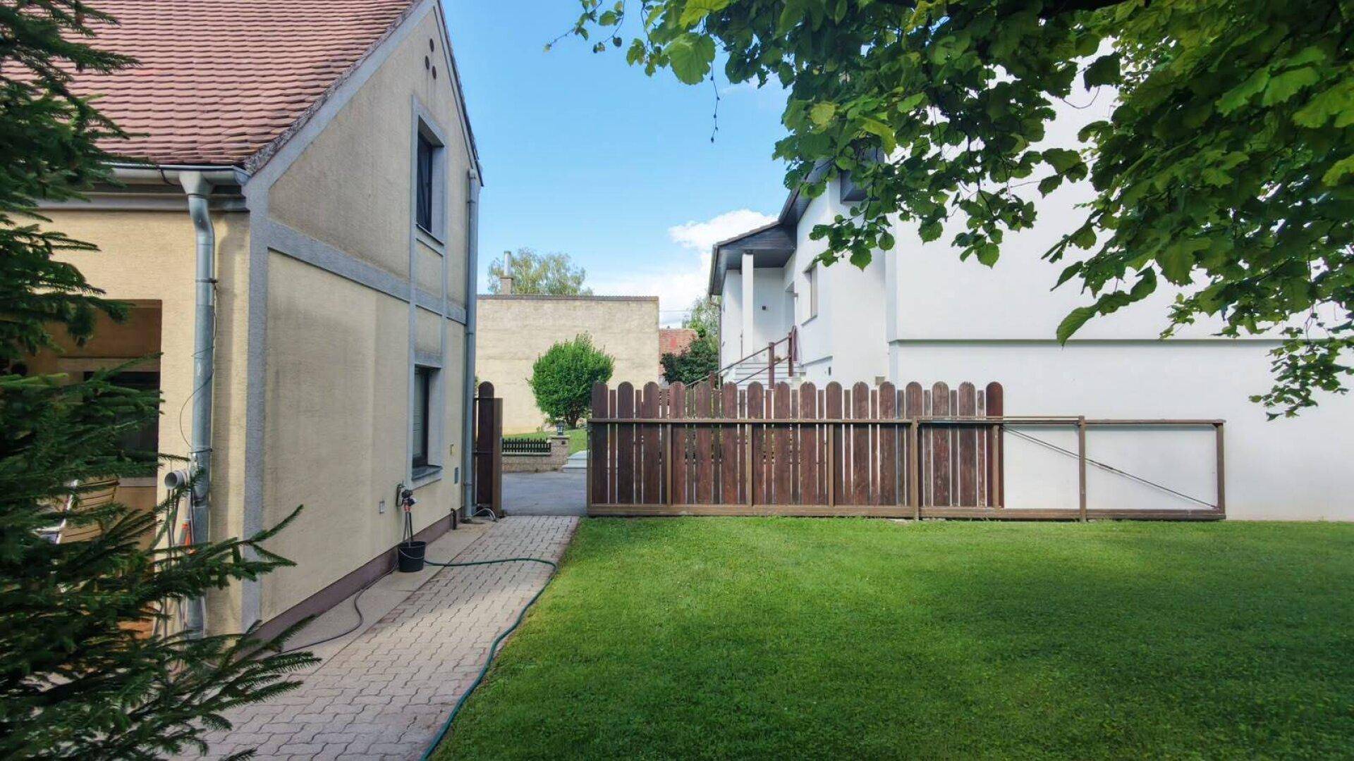 Gepflasterter Gartenweg entlang des Hauses mit Rasenfläche und Holzzaun.