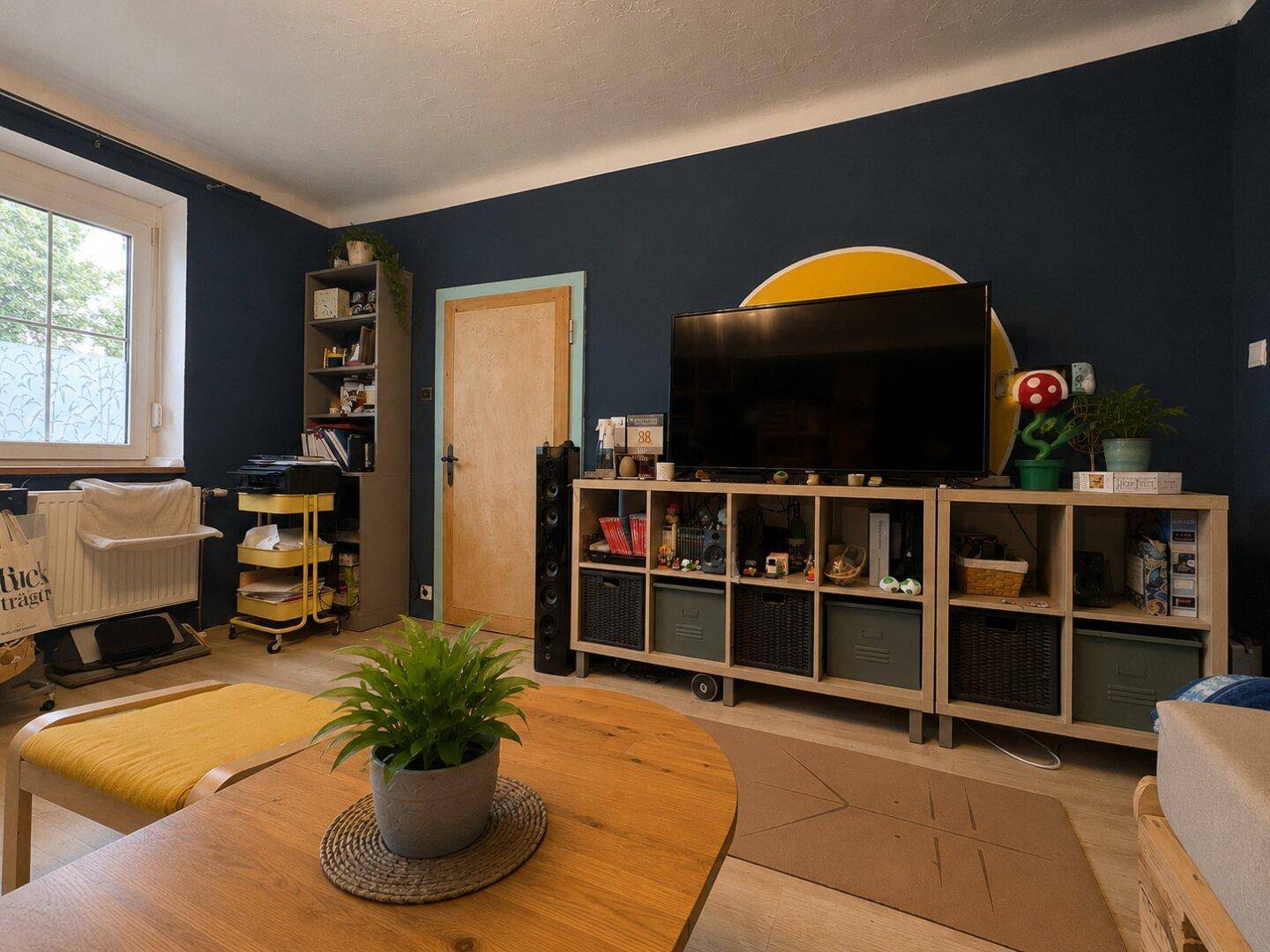 Wohnzimmer mit Fernseher auf einem Regal, großem Fenster und farbenfroher Dekoration.