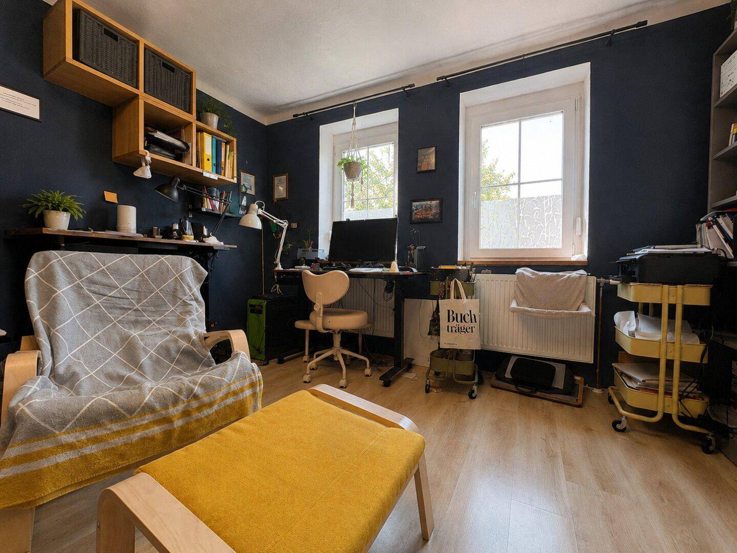 Arbeitszimmer mit dunkelblauen Wänden, zwei Fenstern und einem Schreibtischbereich.