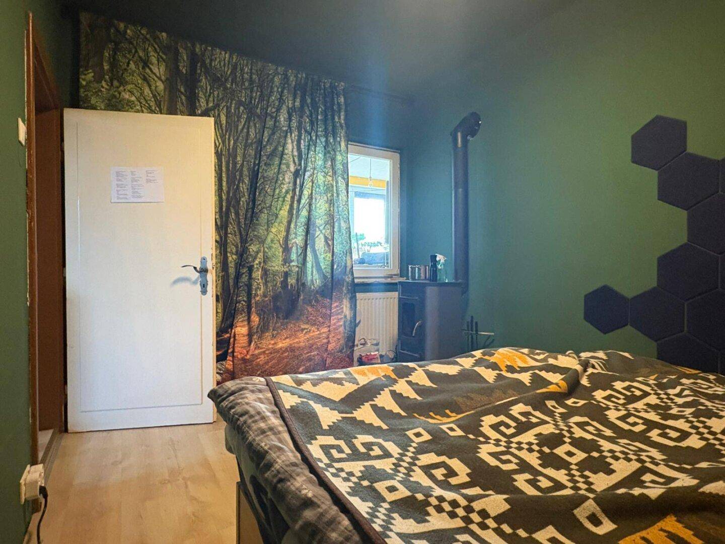 Schlafzimmer mit Kaminofen, Fenster mit Waldmotiv-Vorhang und Holzboden.