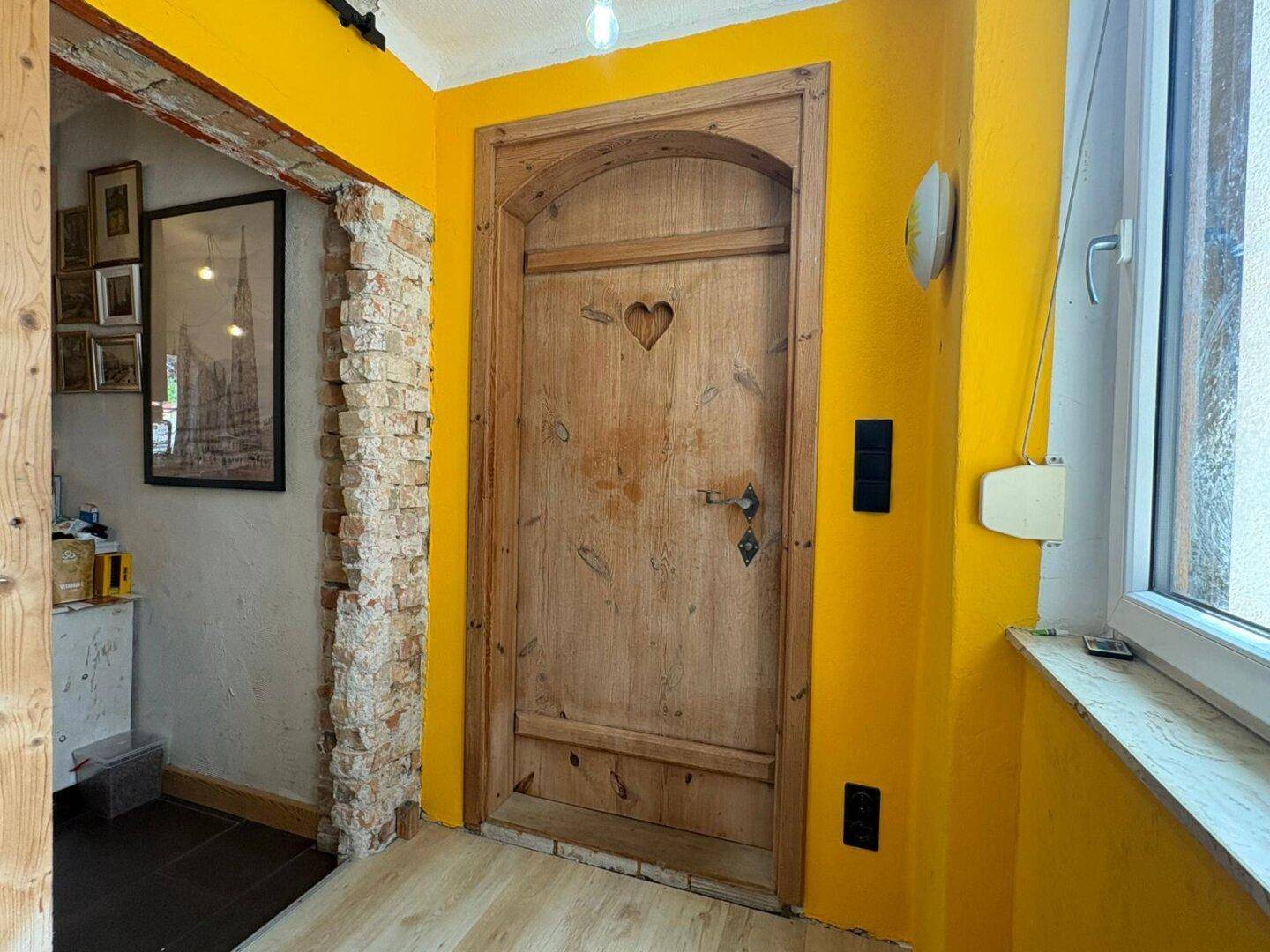 Heller Flur mit leuchtend gelben Wänden, einer Holztür mit Herzdetail und freigelegtem Mauerwerk.