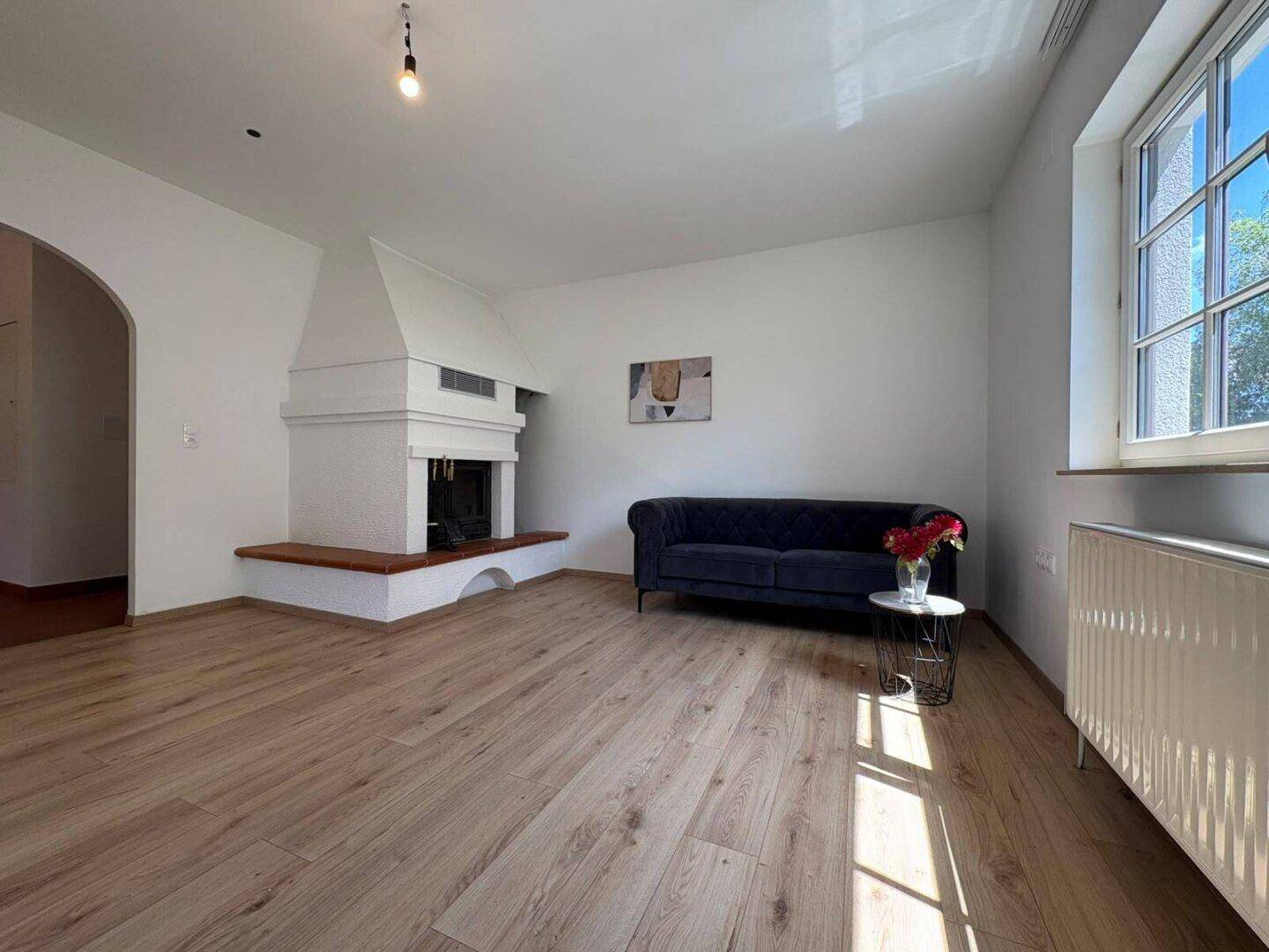 Helles Wohnzimmer mit gemütlichem Sofa, großem Kamin und viel Tageslicht durch das Fenster.