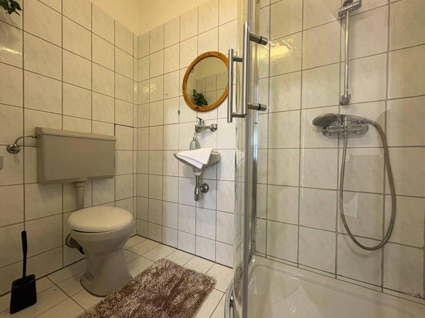Kleines Badezimmer mit Dusche, WC und Waschbecken, komplett gefliest in Weiß.
