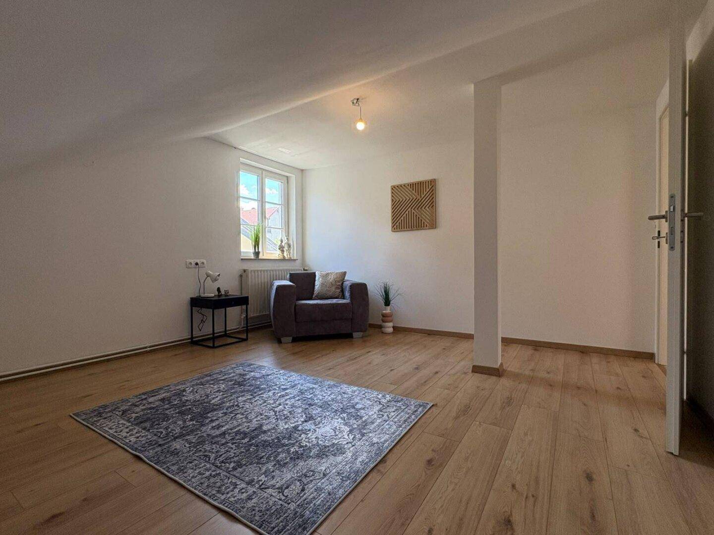 Helles Zimmer im Dachgeschoss mit Fenster, Sessel und Holzboden, unter schräger Decke.