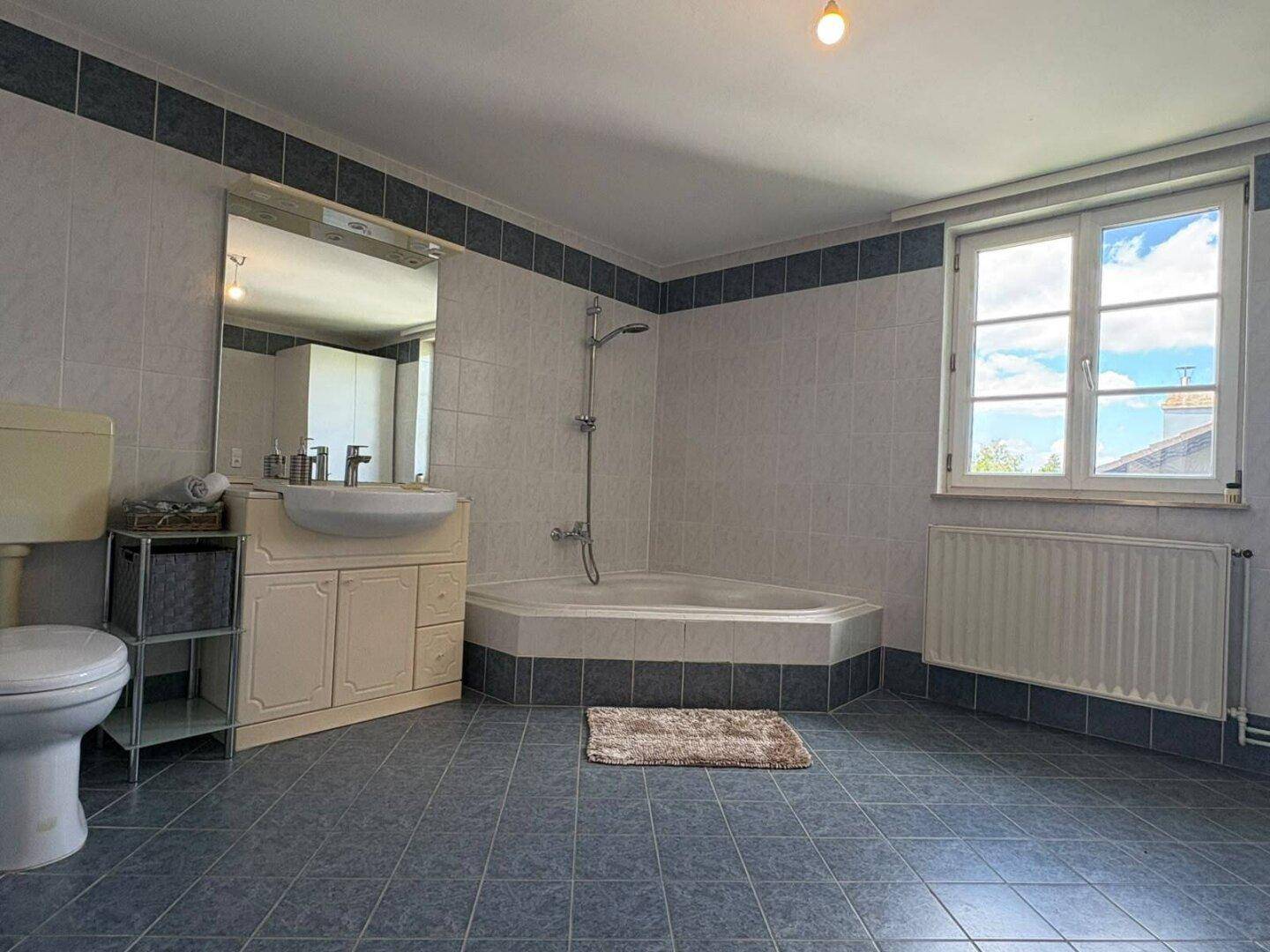 Großes Badezimmer mit Eckbadewanne, WC, Waschbecken und Fenster, gefliest in Blau und Weiß.
