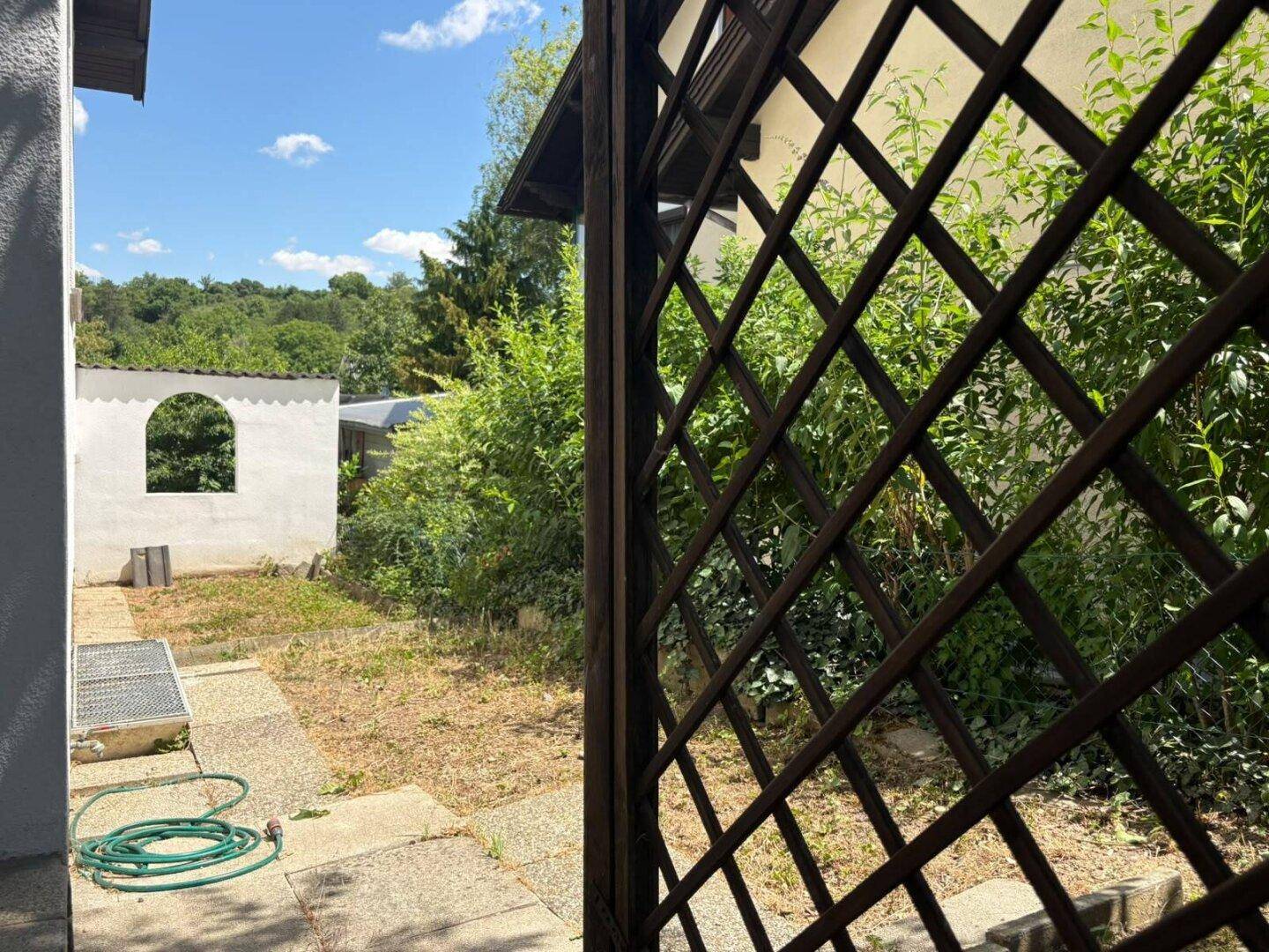 Blick in den Gartenbereich durch ein Holzgitter, mit viel Grün und einem kleinen Schuppen.