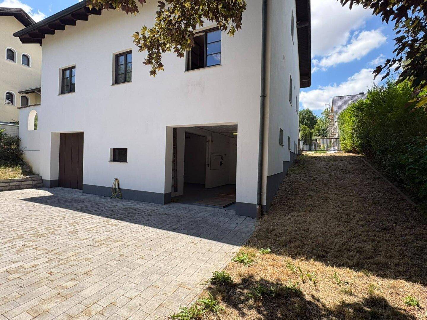 Seitenansicht des weißen Gebäudes mit Garagenzugang, gepflasterter Auffahrt und einem grasbewachsenen Hang.