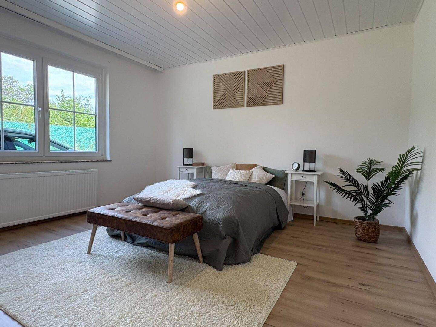 Gemütliches Schlafzimmer mit Laminatboden, weißer Holzdecke und großem Fenster mit Blick ins Grüne.