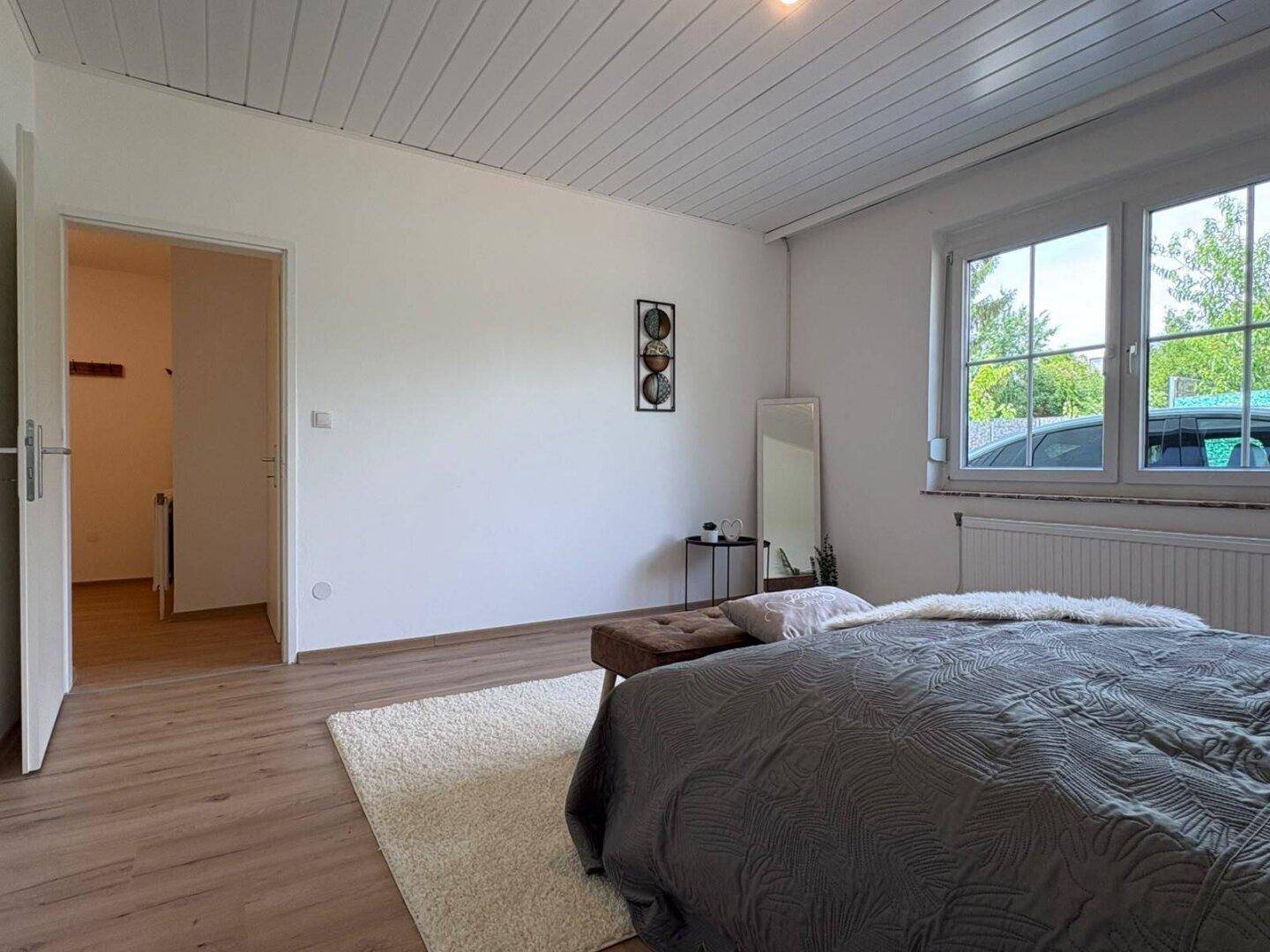 Helles Schlafzimmer mit Laminatboden, weißer Holzdecke und einem großen Fenster, das viel Licht hereinlässt.